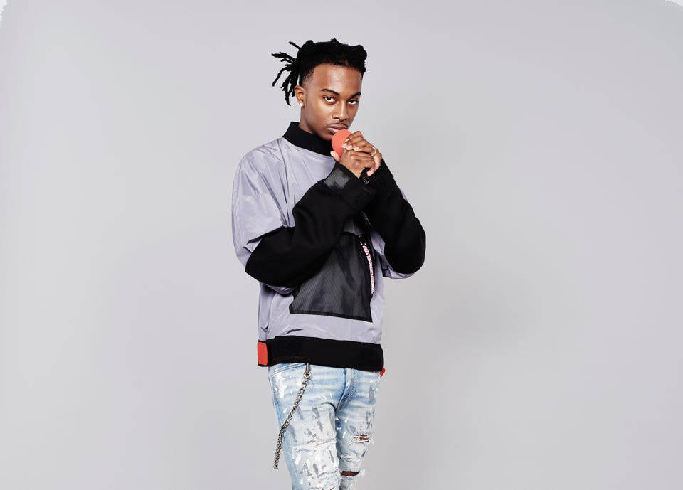 Carti 桌布 壁紙 — 一位時尚的年輕男子留著辮子，穿著灰色和黑色層次夾克和破洞牛仔褲，在純灰色背景前自信地擺姿勢。