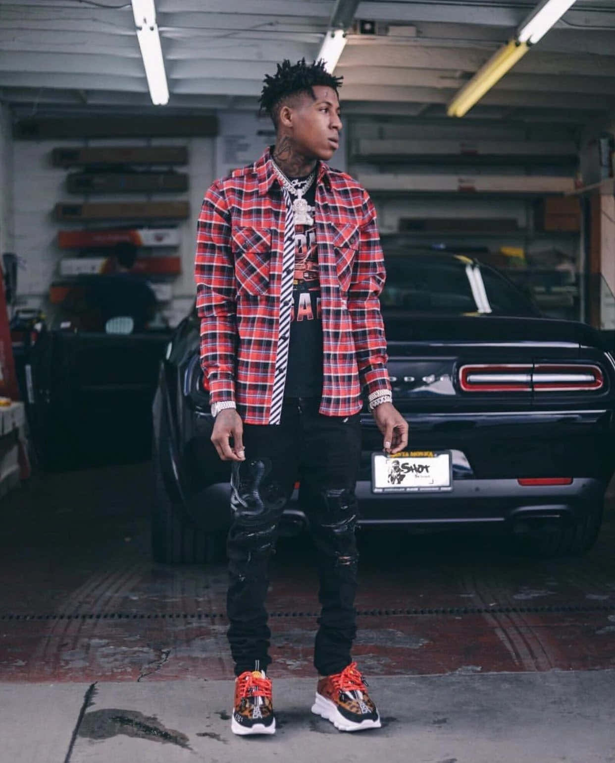 NBA YoungBoy 桌布 壁紙 — 一位時尚的年輕男子穿著紅色格紋襯衫和破洞牛仔褲，自信地站在車庫裡，旁邊是一輛黑色的道奇挑戰者。