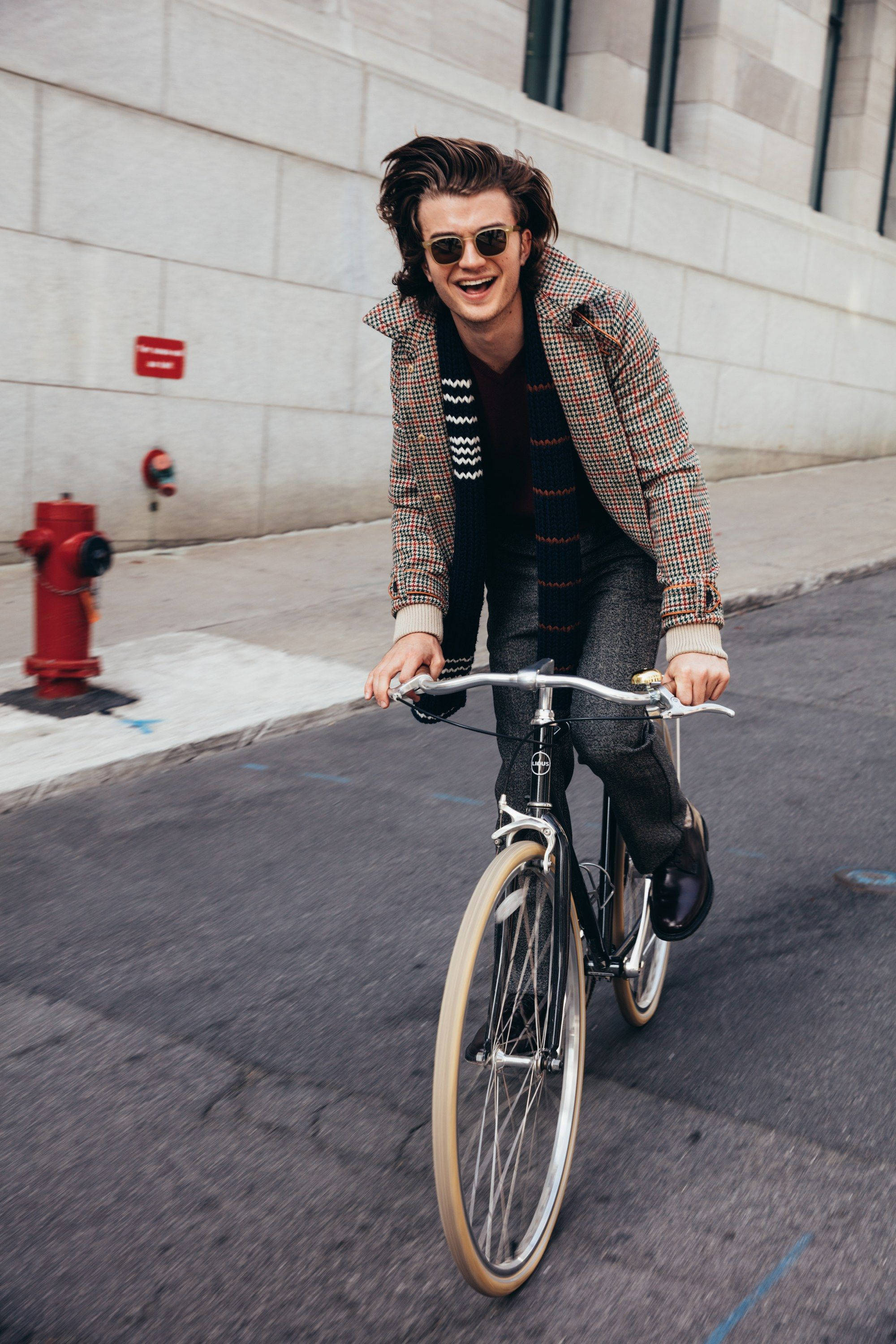 Joe Keery 桌布 壁紙 — 一位時尚的年輕男子笑著騎著復古自行車沿著城市人行道經過一個紅色消防栓。