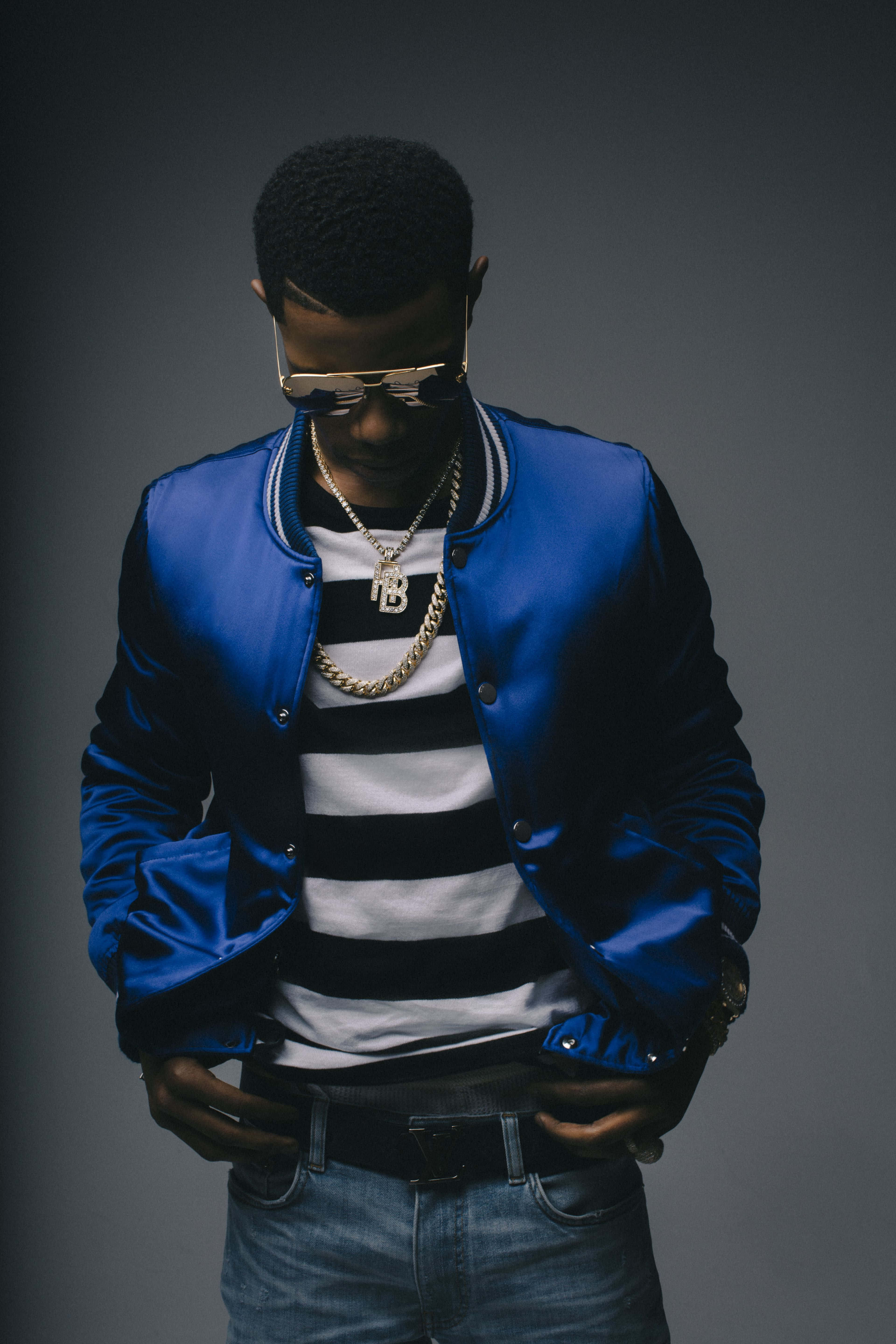 Rappers 桌布 壁紙 — 一位時尚的年輕男子身穿閃亮藍色飛行員夾克和金鏈，在深灰色工作室背景前自信地擺姿勢。