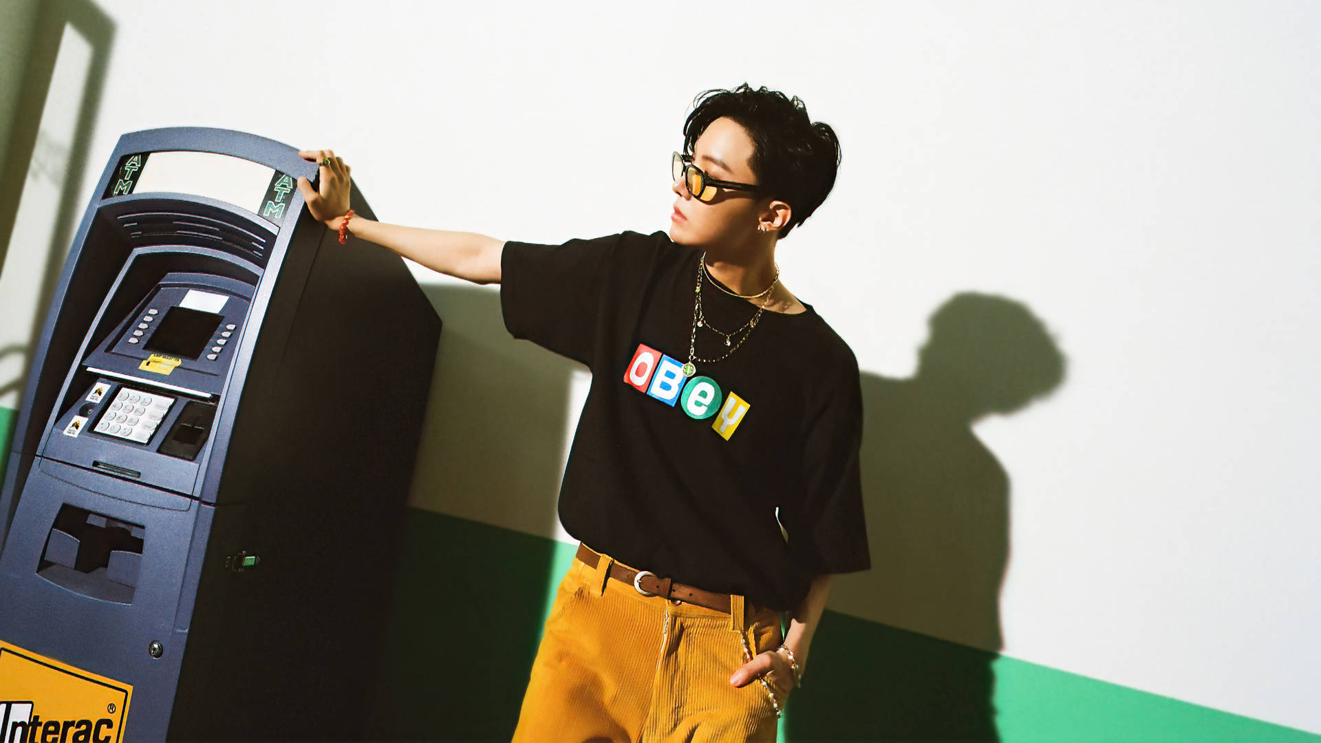 jhope 桌布 壁紙 — 一位時尚的年輕男子身穿黑色OBEY T恤和芥末色褲子，靠在一部藍色提款機上，在白色和綠色的牆上投下清晰的陰影。