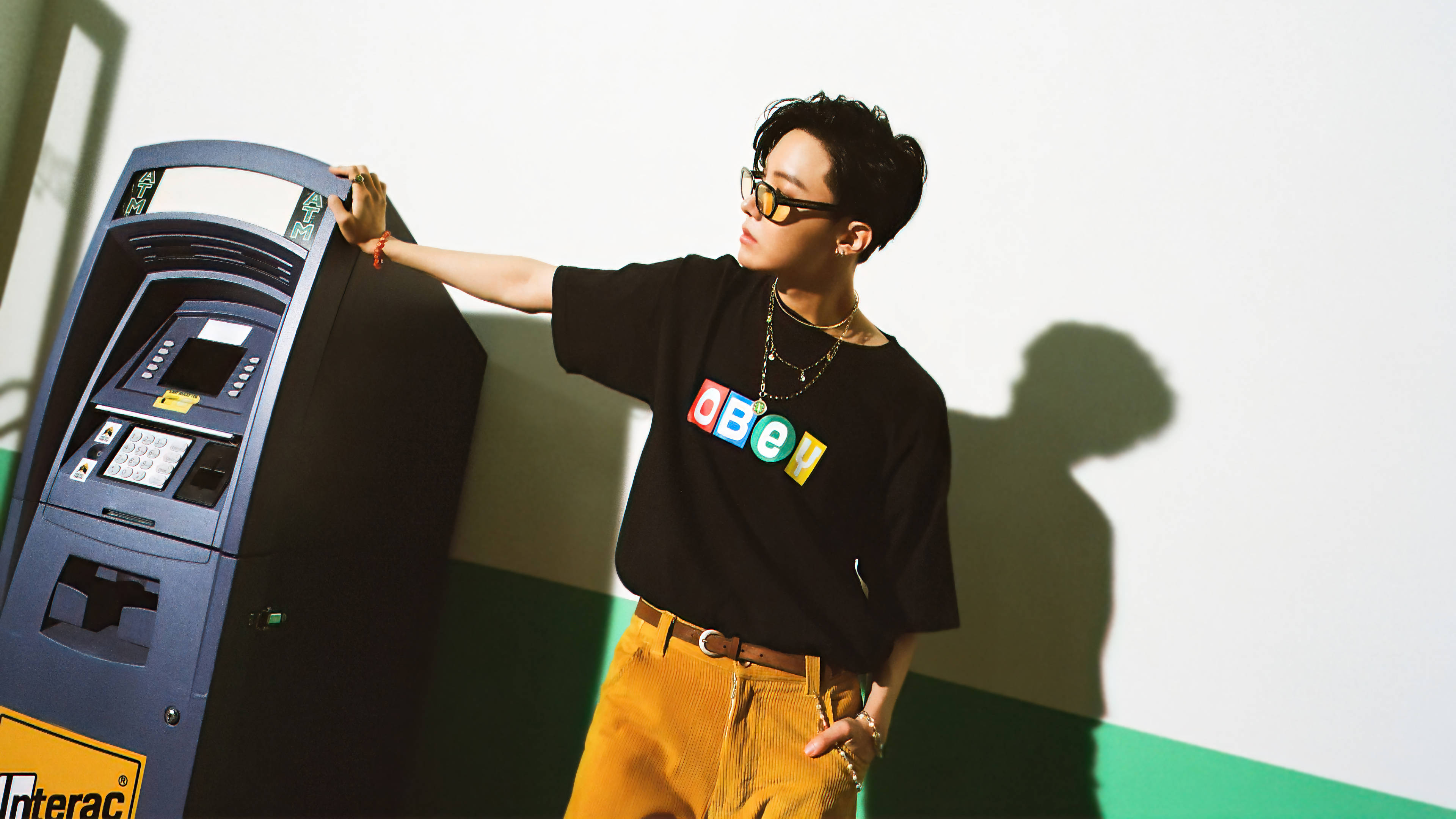 j-hope 桌布 壁紙 — 一位時尚的年輕男子身穿黑色OBEY T恤和芥末黃褲子，靠在一台藍色自動櫃員機上，在白綠相間的牆上投下大膽的影子。
