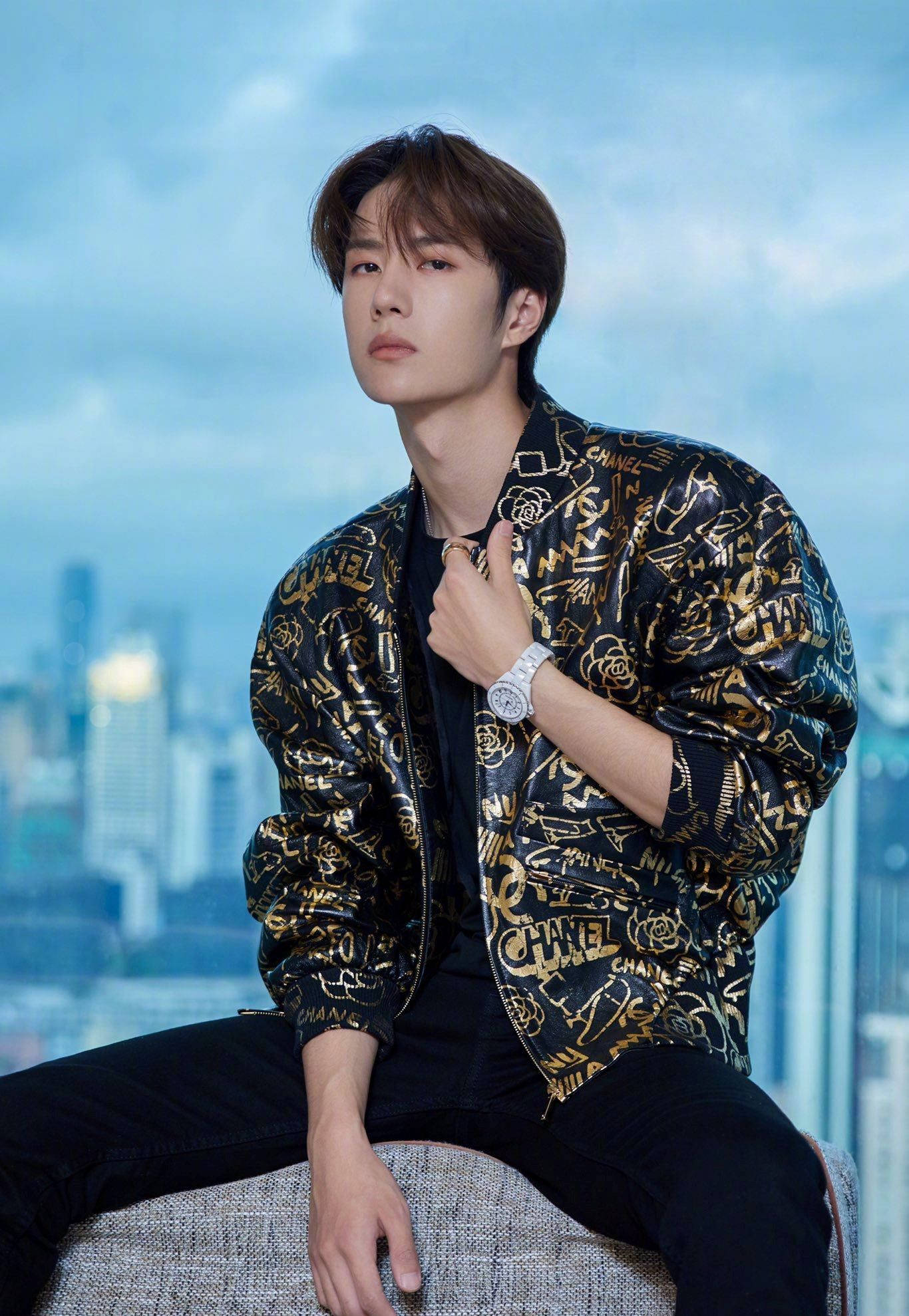 Wang Yibo 桌布 壁紙 — 一位時尚的年輕男子身穿黑金色Chanel飛行員夾克，自信地坐在一張有紋理的沙發上，背景是模糊的城市天際線。