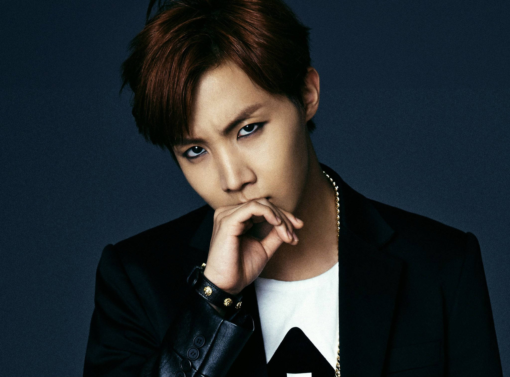 j-hope 桌布 壁紙 — 一位時尚的年輕紅髮男子，眼神銳利，穿著黑色西裝外套和金鏈，在深藍色工作室背景前擺姿勢。