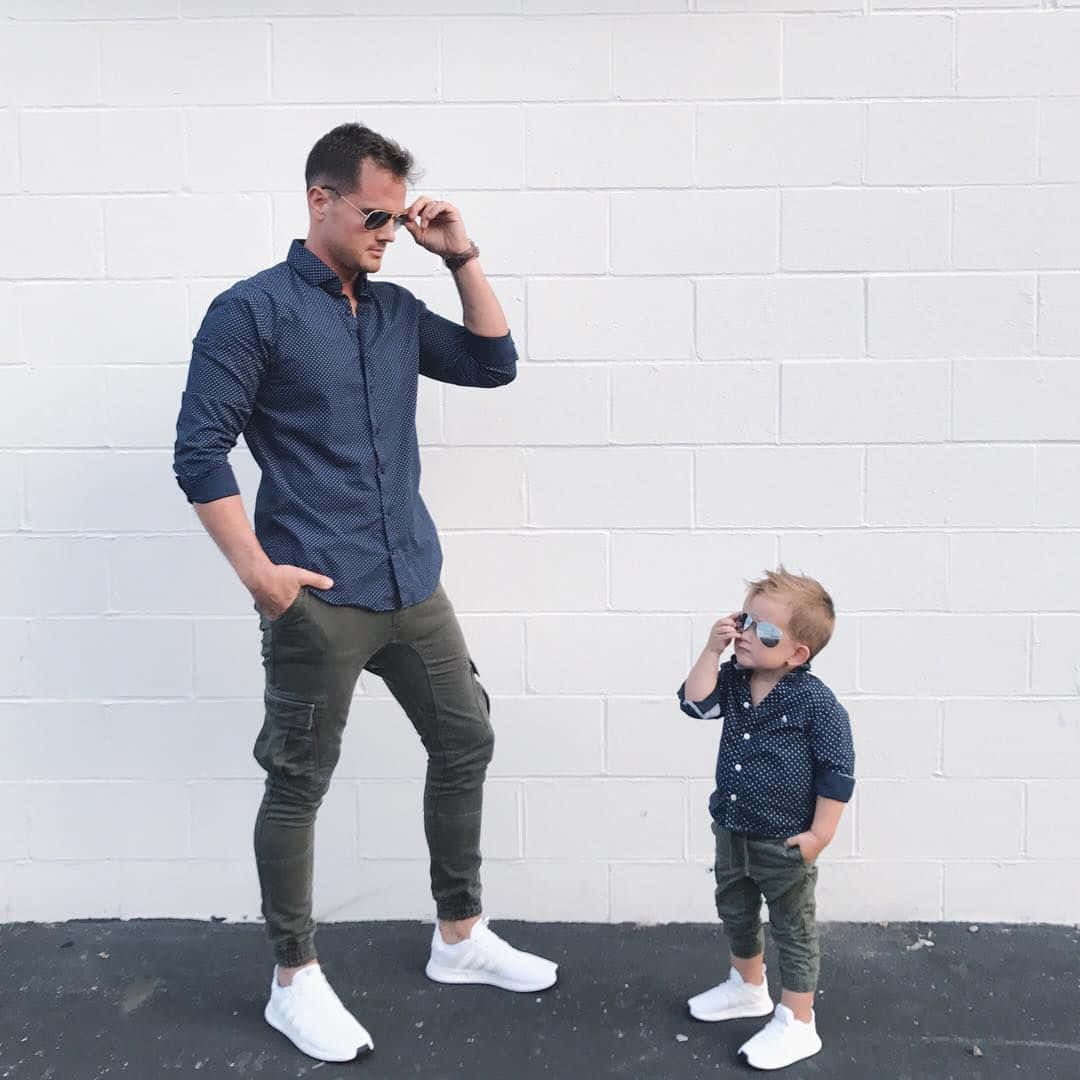Father And Son 桌布 壁紙 — 一位時尚的成人和幼兒穿著相配的藍色圖案襯衫和太陽眼鏡，自信地靠在白色磚牆上擺姿勢。