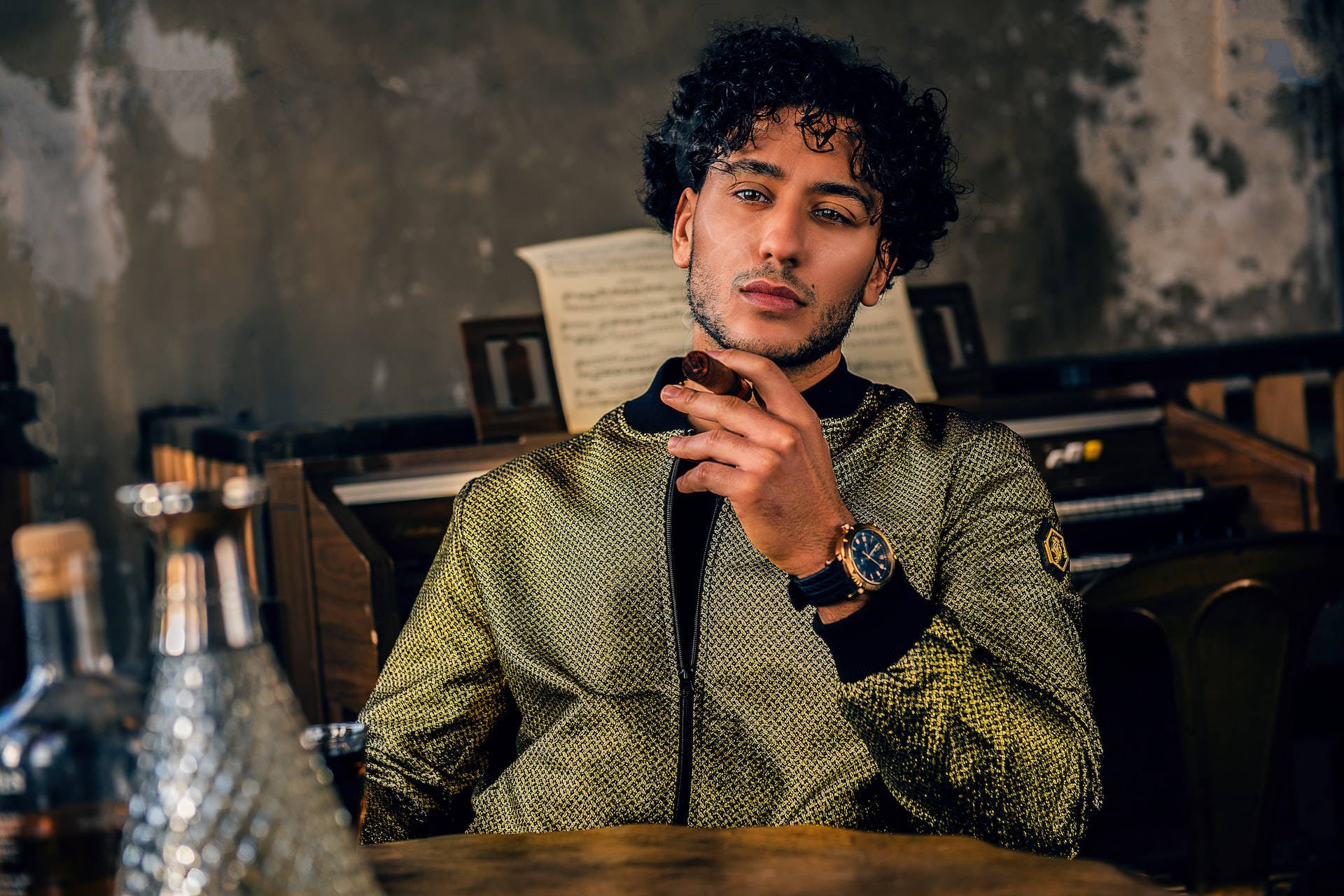 Curly Hair 桌布 壁紙 — 一位時尚的捲髮男士坐在木桌旁，在光線昏暗的鄉村風格房間內手持雪茄，房間帶有復古裝飾。