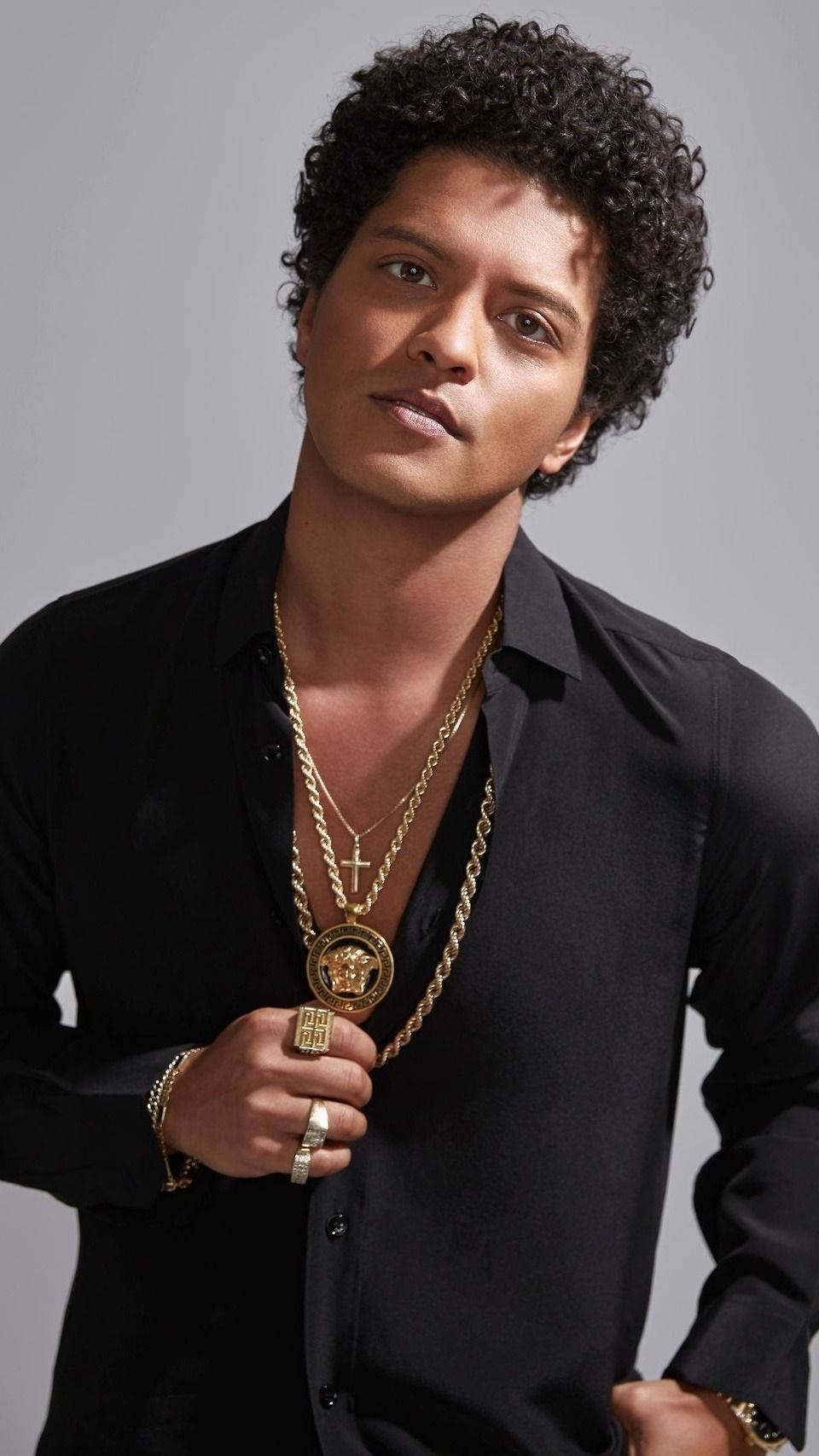 Bruno Mars 桌布 壁紙 — 一位時尚的捲髮男士穿著黑色襯衫，佩戴多層金鍊，背景為中性灰色。