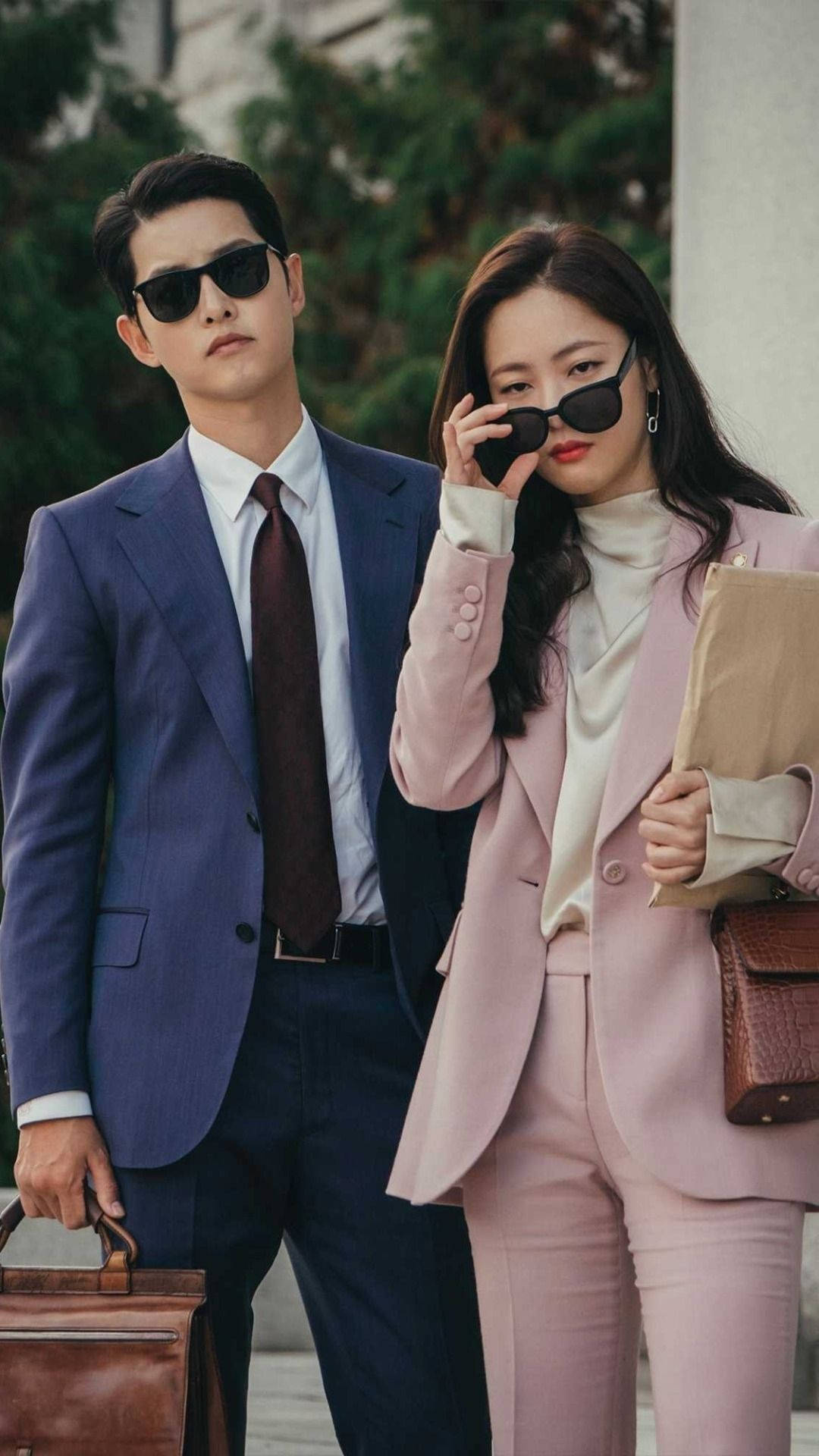 文森佐 桌布 壁紙 — 一位時尚的男士和女士穿著訂製西裝和太陽眼鏡，自信地在戶外拿著公事包擺姿勢。