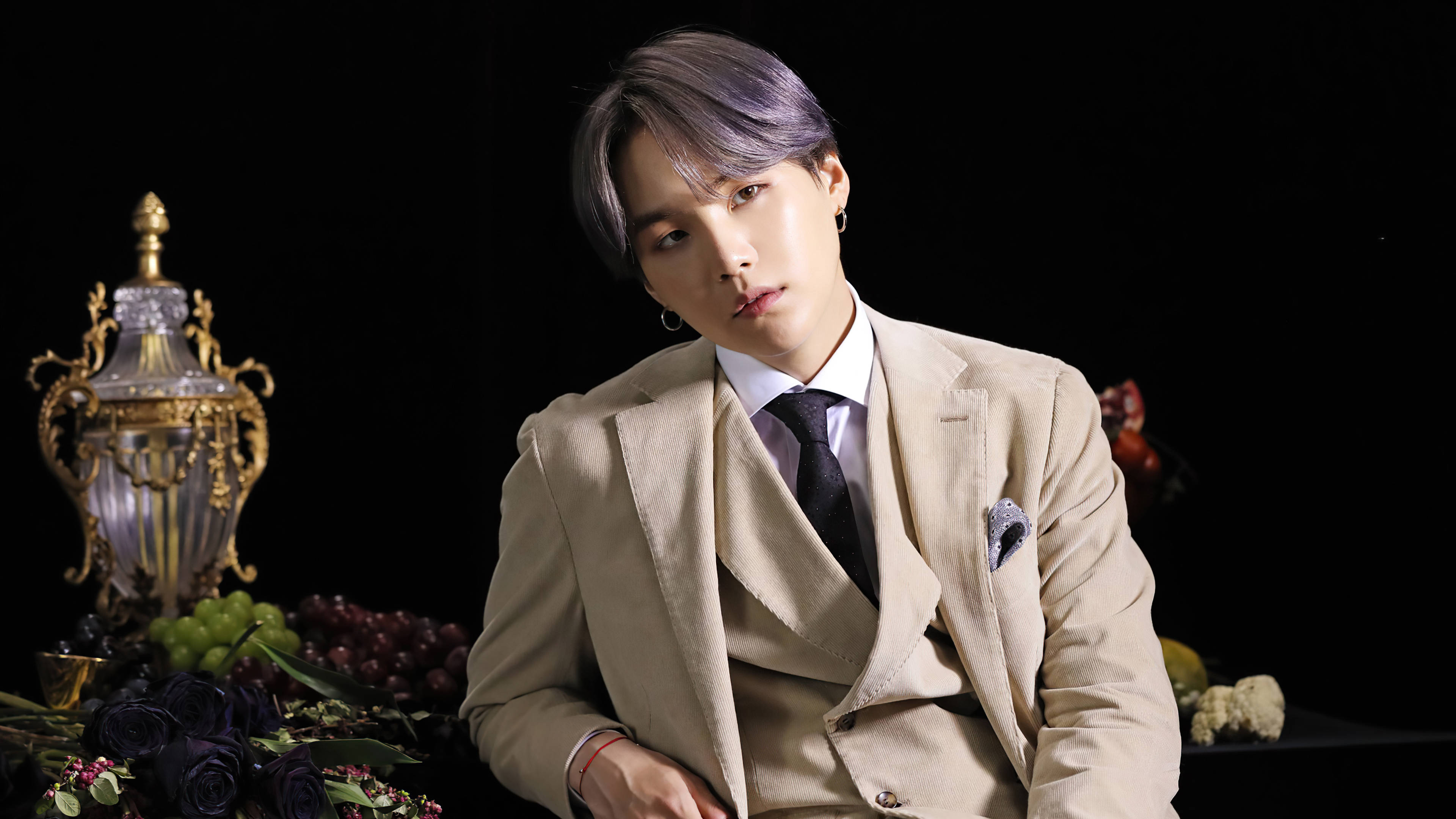 슈가 桌布 壁紙 — 一位時尚的紫髮男士穿著米色西裝，背景為黑色，配有裝飾花瓶、葡萄和深色玫瑰。
