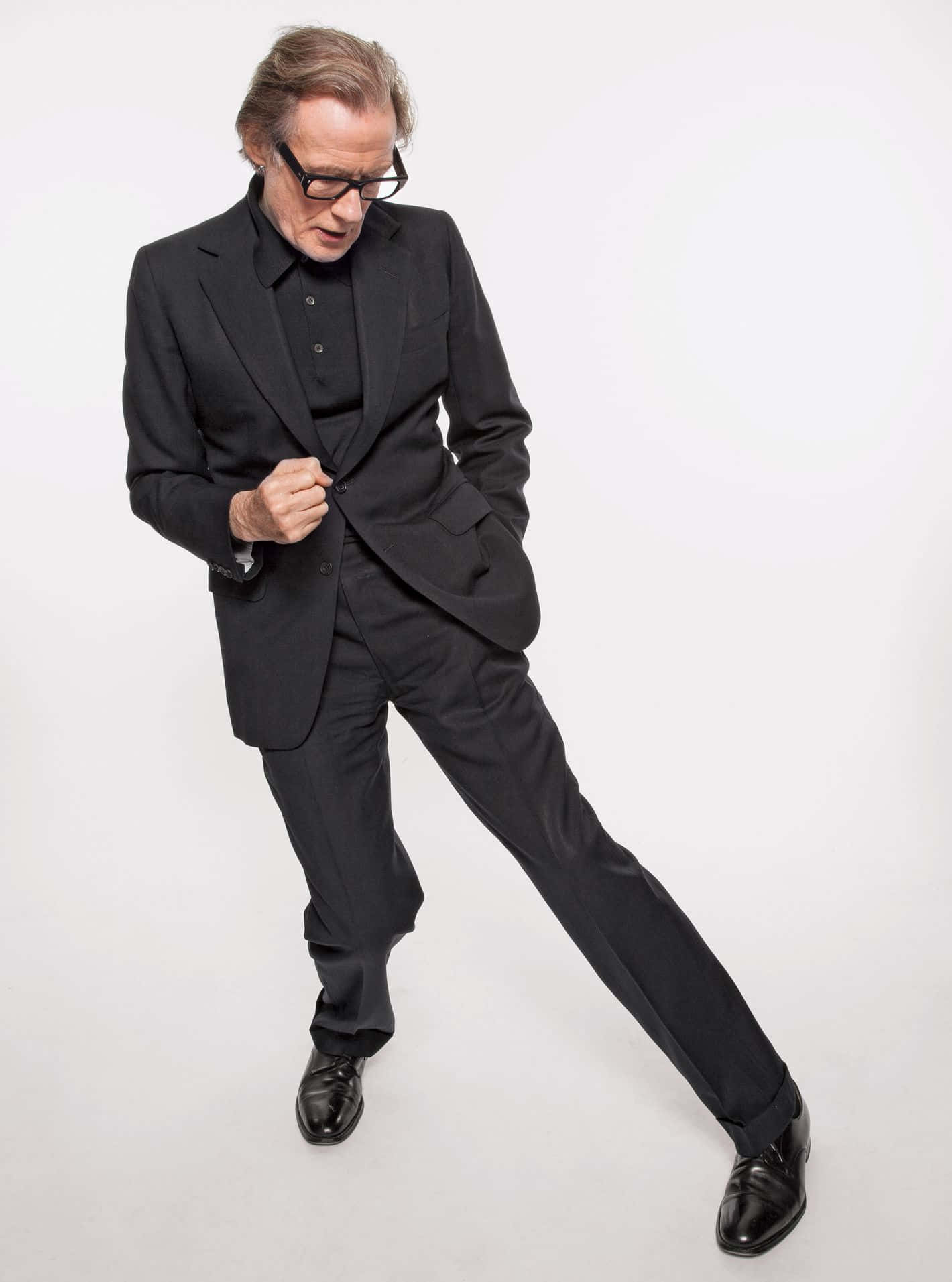 Man In Suit 桌布 壁紙 — 一位時尚的老年男士穿著黑色西裝和眼鏡，在純白背景前擺出動態姿勢。