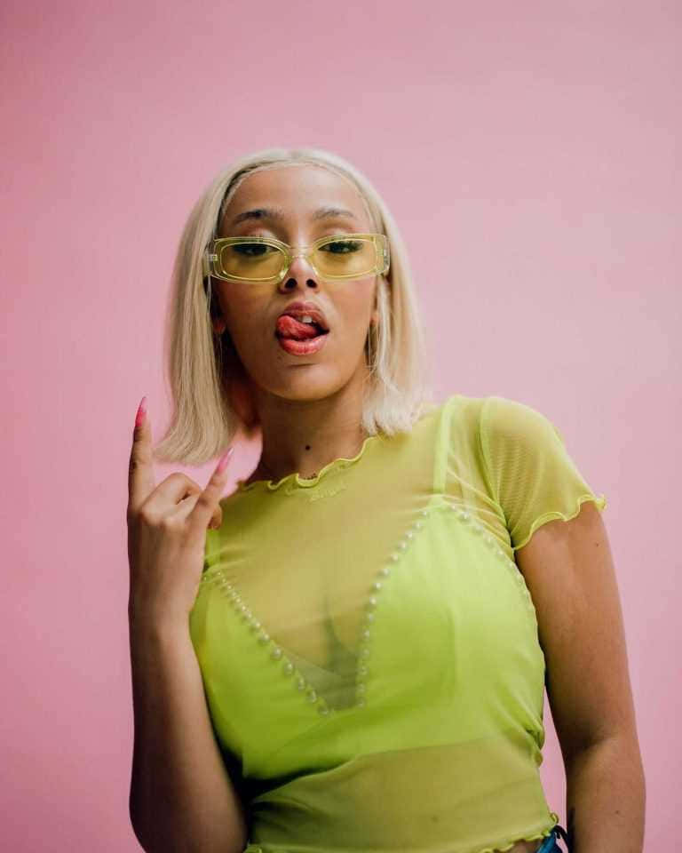 Doja Cat 桌布 壁紙 — 一位時尚的金髮女性戴著黃色眼鏡，在純粉紅色背景上頑皮地伸出舌頭。