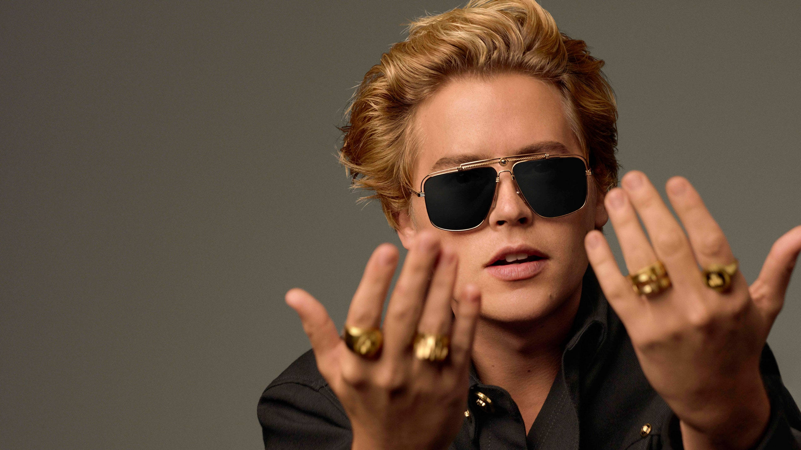Sunglasses 桌布 壁紙 — 一位時尚的金髮男子戴著飛行員太陽眼鏡，雙手戴著金戒指，在灰色中性背景前擺姿勢。