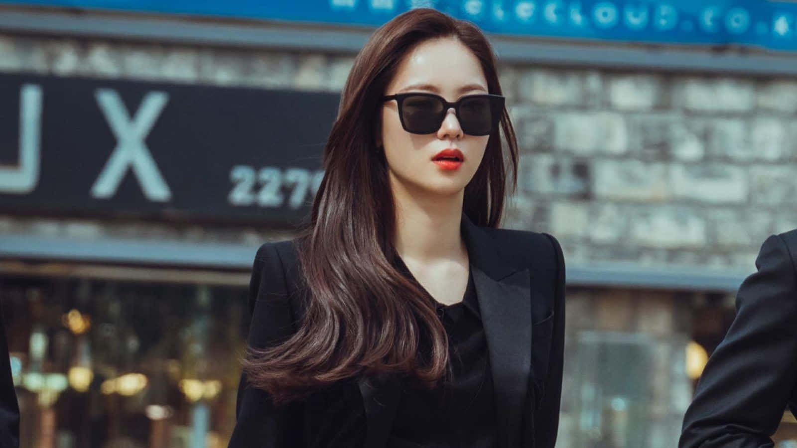 Beautiful Fashion 桌布 壁紙 — 一位時尚的長棕髮女性戴著黑色太陽眼鏡和一件俐落的黑色西裝外套，走在城市街道上。