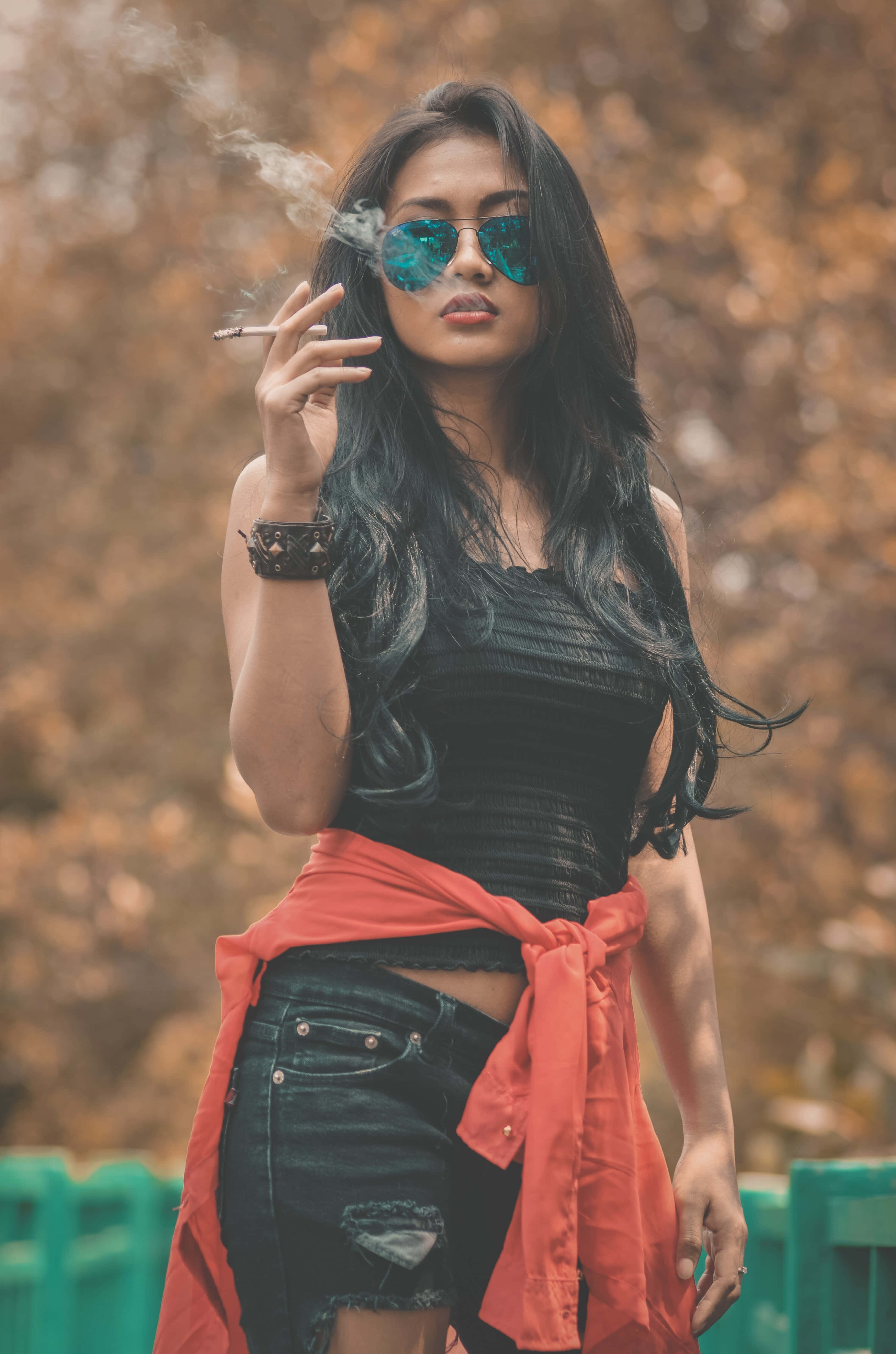 Woman 桌布 壁紙 — 一位時尚的長黑髮女性，戴著反光太陽眼鏡，紅色外套綁在腰間，在模糊的秋日背景下抽煙。