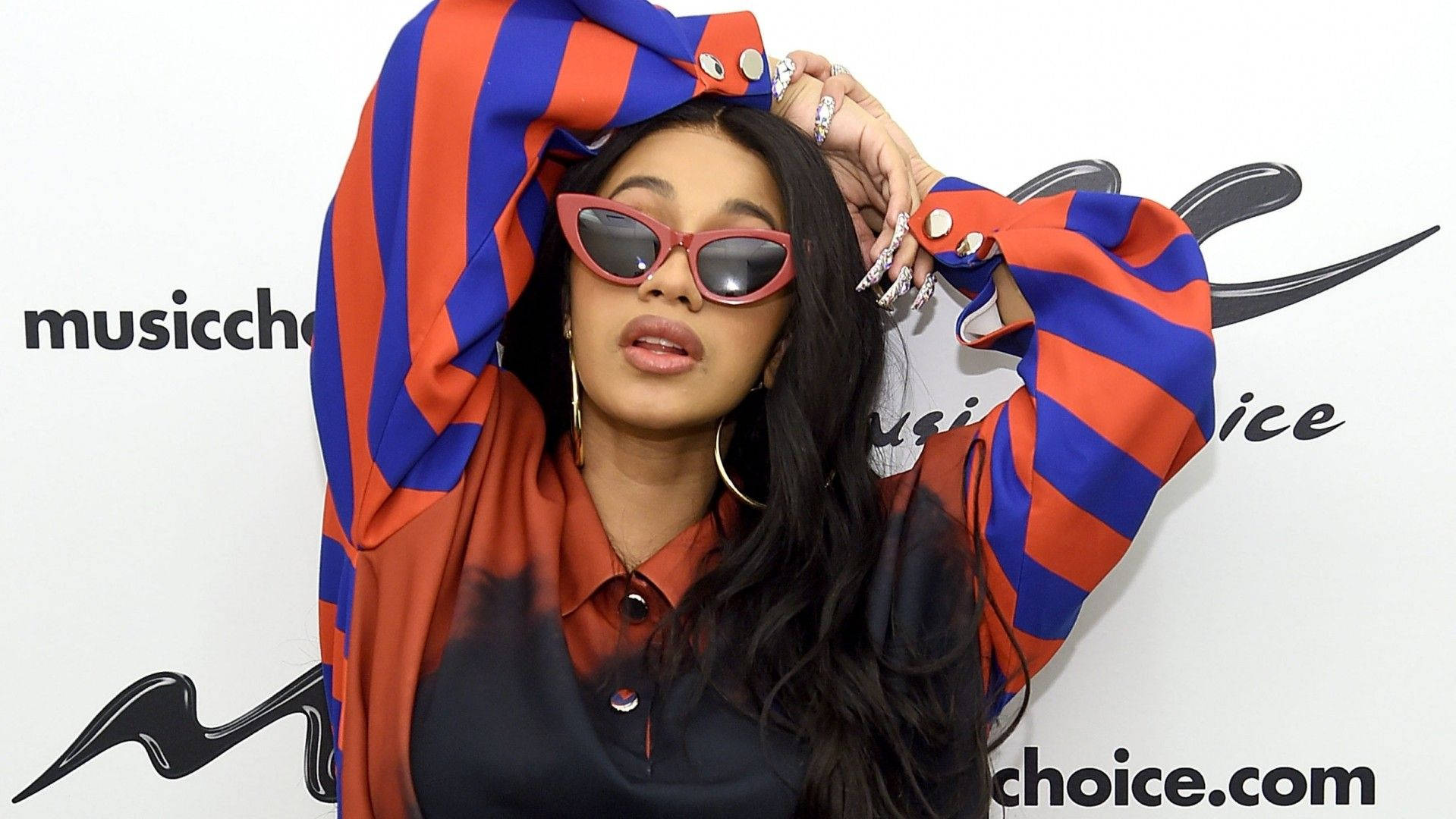 cardi 桌布 壁紙 — 一位時尚的長黑髮女性，穿著大膽的紅藍條紋袖子和粉紅色貓眼太陽眼鏡，在白色背景前擺姿勢，背景上有黑色Music Choice標誌。