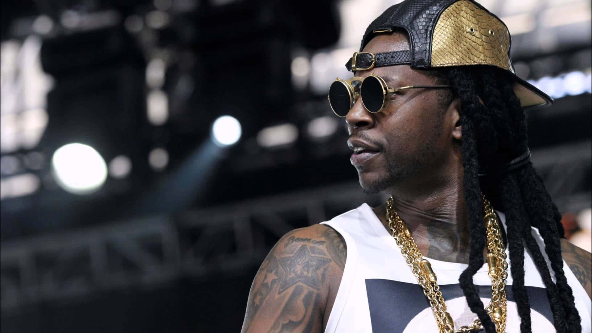 2 Chainz 桌布 壁紙 — 一位時尚的饒舌歌手，留著辮子頭，戴著金鍊，自信地站在燈光昏暗的演唱會舞台上。