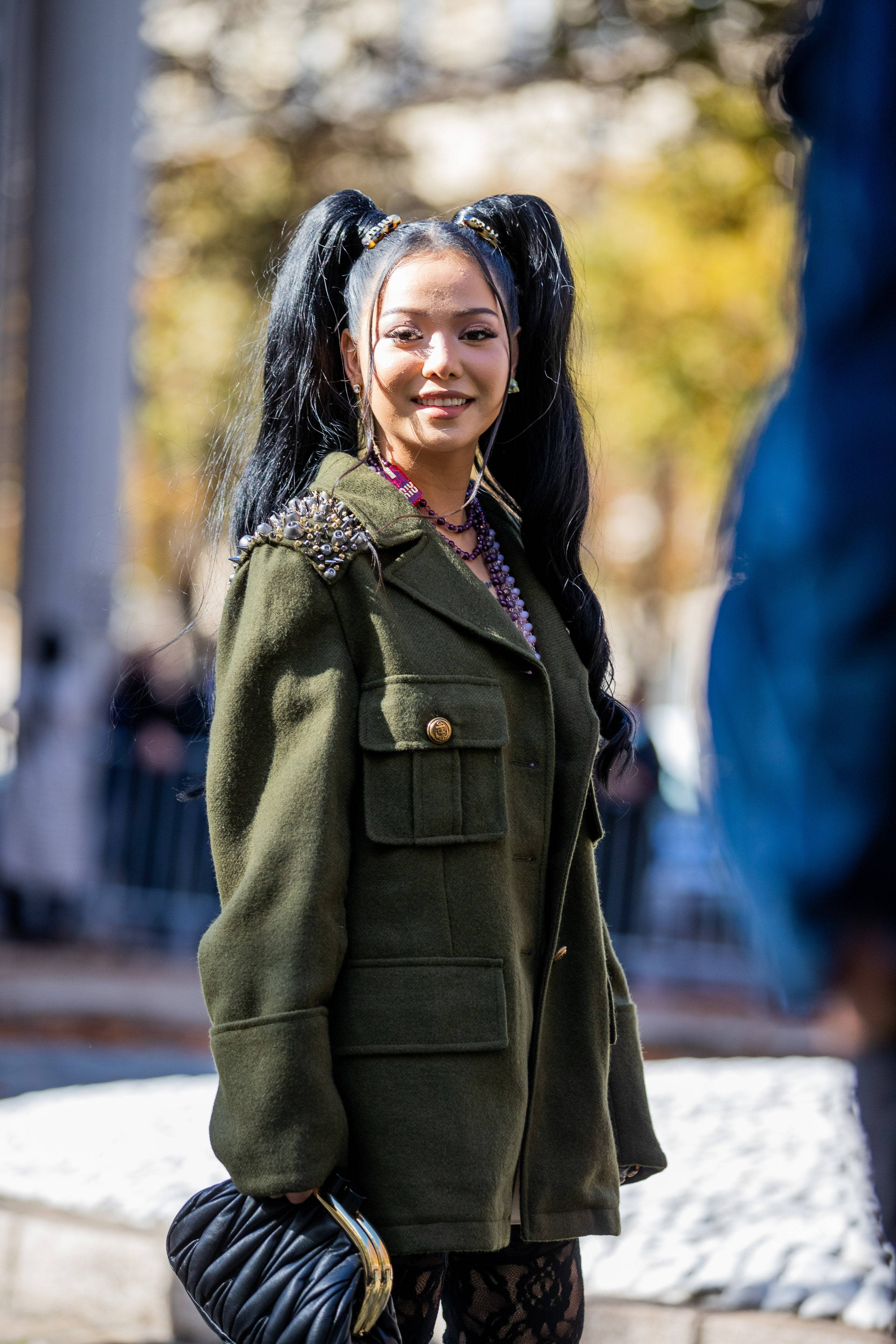 Bella Poarch 桌布 壁紙 — 一位時尚的黑辮子女性微笑著，身穿飾有鉚釘的橄欖綠軍裝外套，手持黑色絎縫手拿包。