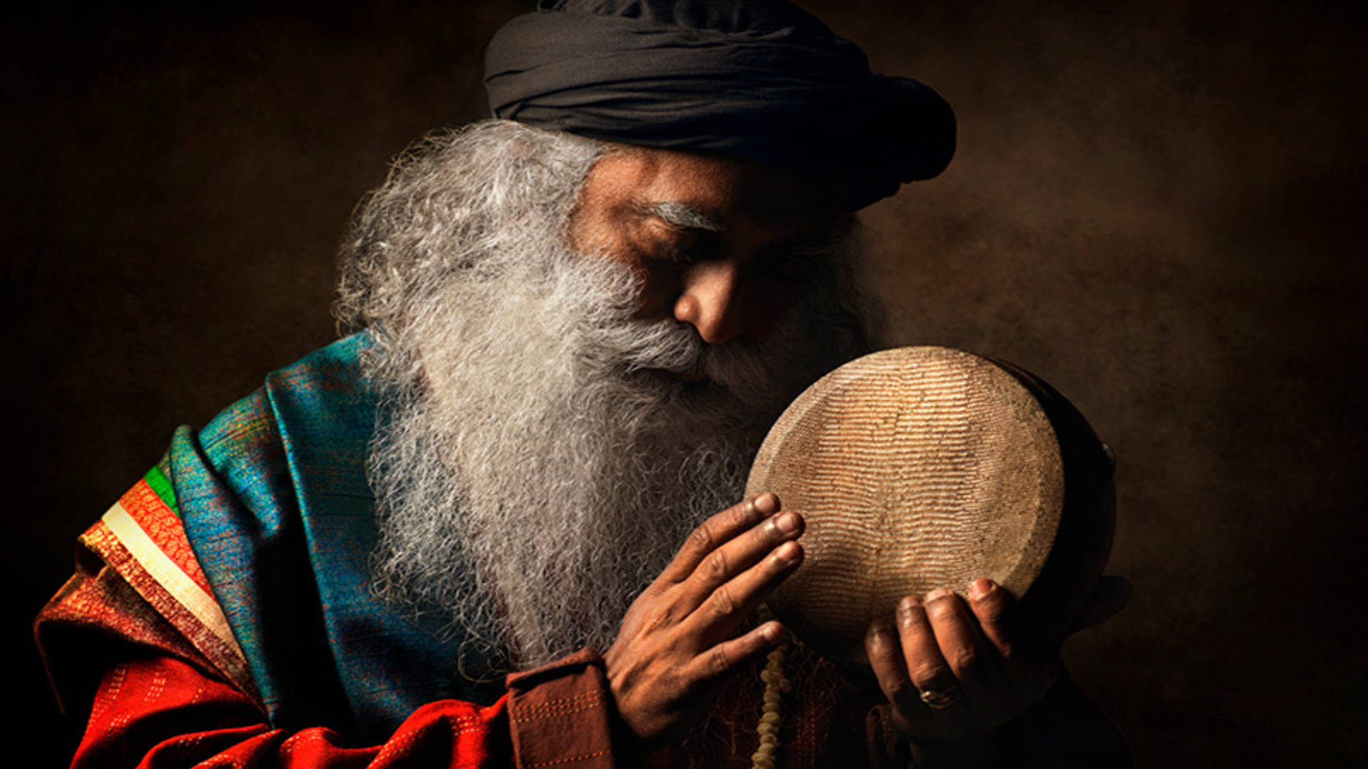 Sadhguru 桌布 壁紙 — 一位智慧長者留著長長的白鬍子，在燈光昏暗的工作室中，在戲劇性的燈光下拿著手鼓。