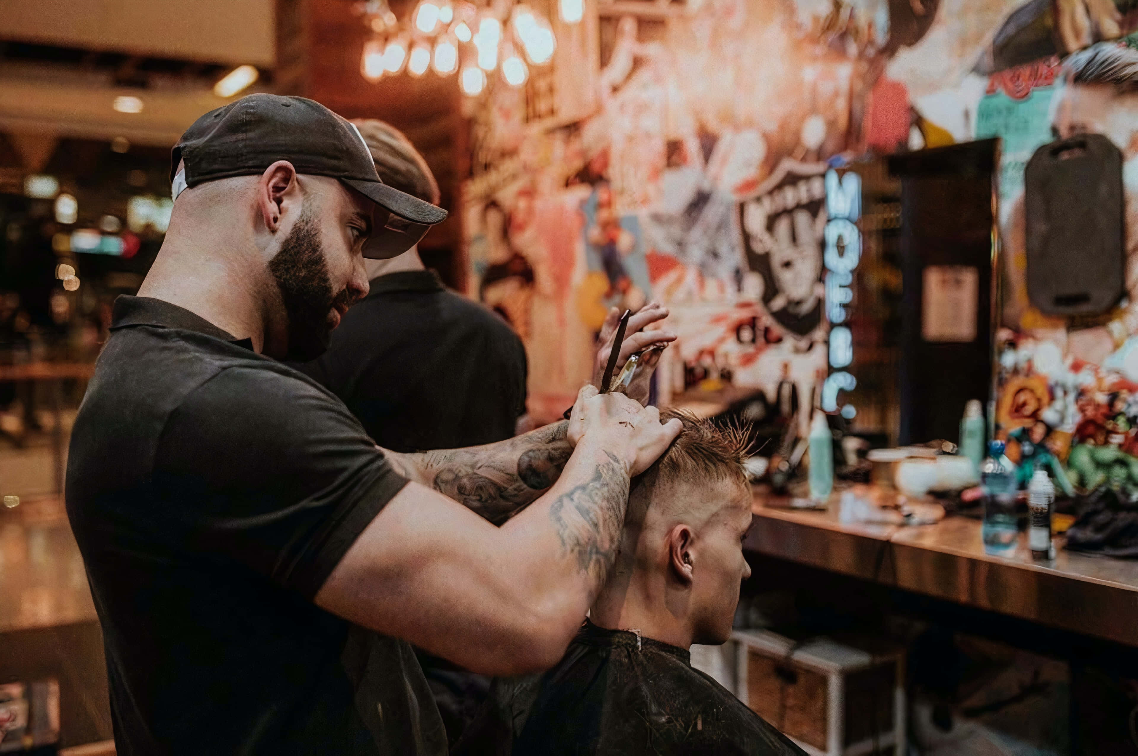 Barber 桌布 壁紙 — 一位有刺青、戴帽子的理髮師在燈光溫暖、牆上有塗鴉的理髮店裡，小心翼翼地為一位年輕男子修剪頭髮。