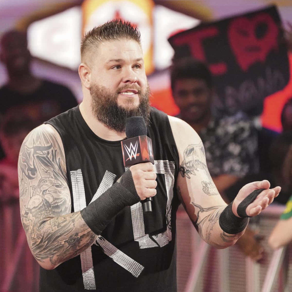 WWE 桌布 壁紙 — 一位有刺青、留鬍子的WWE摔角手在燈光明亮的舞台上對著麥克風說話，身後有模糊的粉絲和標誌。