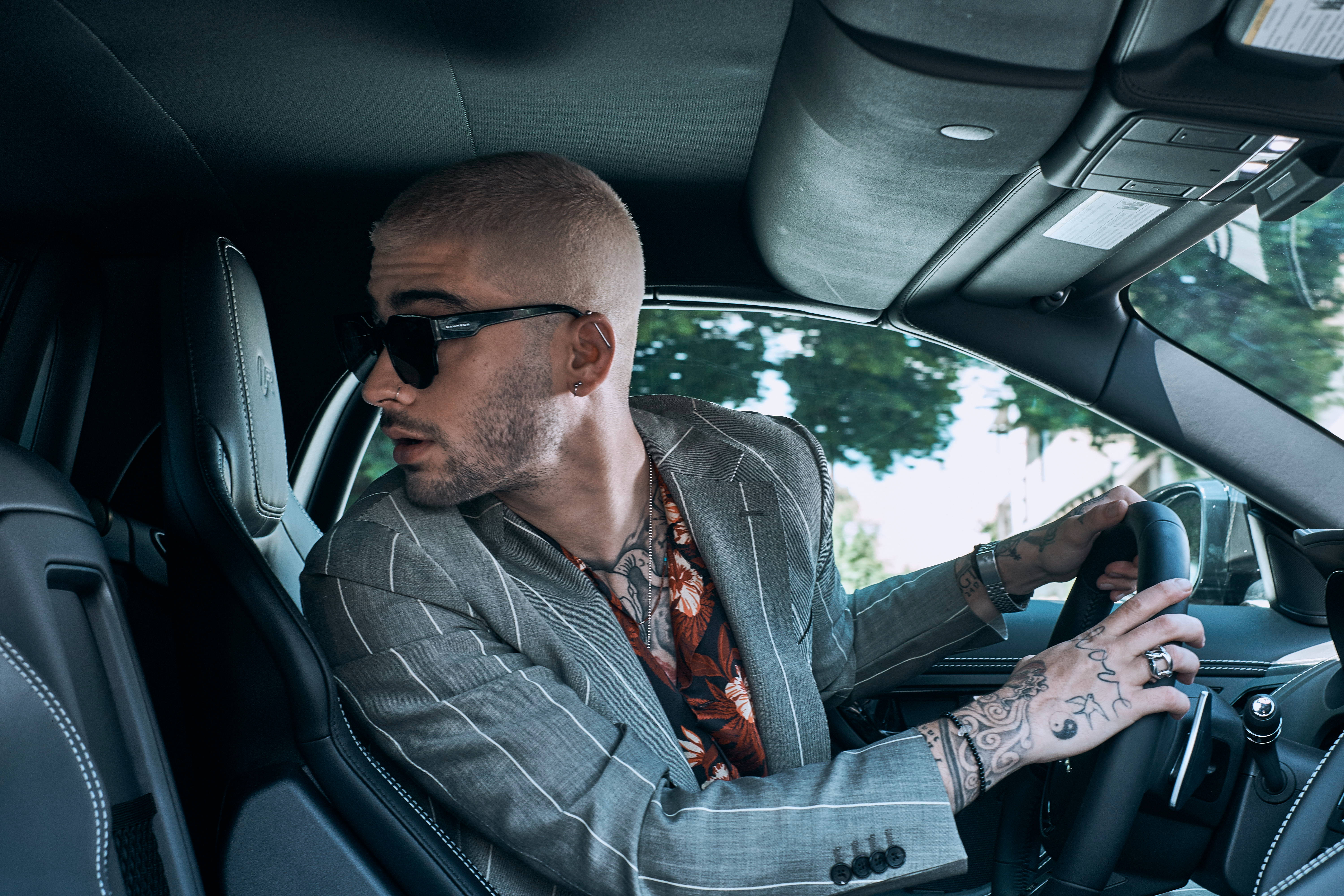 zayn 桌布 壁紙 — 一位有刺青和太陽眼鏡的時尚男士駕駛著一輛皮革內裝的豪華汽車，外面有陽光照射的樹木。