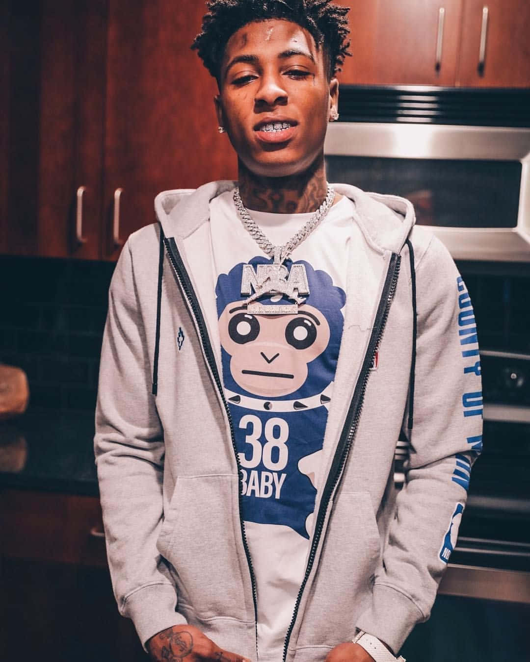 NBA YoungBoy 桌布 壁紙 — 一位有刺青和戴鏈條項鍊的年輕男子站在廚房裡，穿著灰色拉鍊連帽衫，內搭印有卡通猴子的圖案T恤。