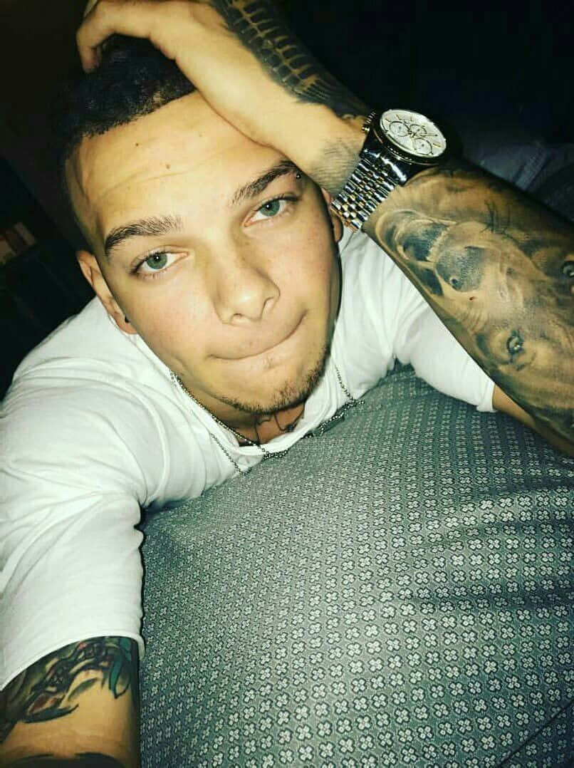 Kane Brown 桌布 壁紙 — 一位有刺青和手錶的年輕男子將頭靠在手臂上，看著鏡頭。