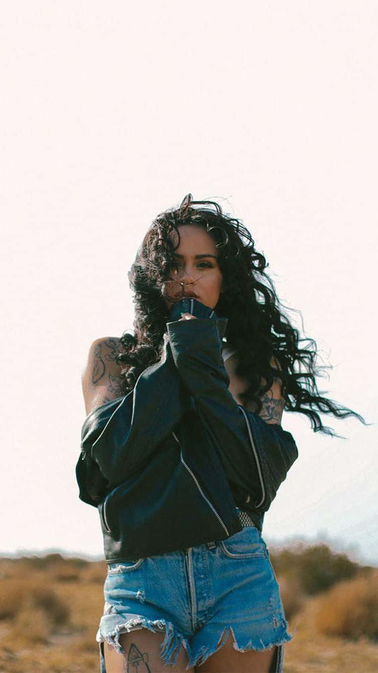 kehlani 桌布 壁紙 — 一位有刺青和捲髮的女性站在沙漠中，穿著黑色外套和破洞牛仔短褲。