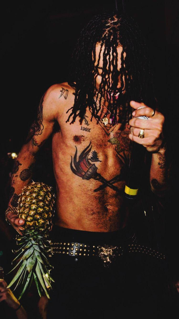 Swae Lee 桌布 壁紙 — 一位有刺青和辮子頭的男人在昏暗的舞台燈光下拿著一個鳳梨和一支麥克風。