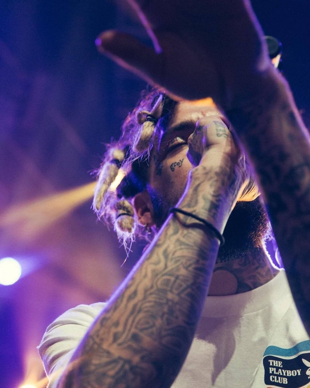 suicideboys 桌布 壁紙 — 一位有刺青和辮子頭的表演者在充滿活力的紫色和黃色舞台燈光下用手遮住臉。