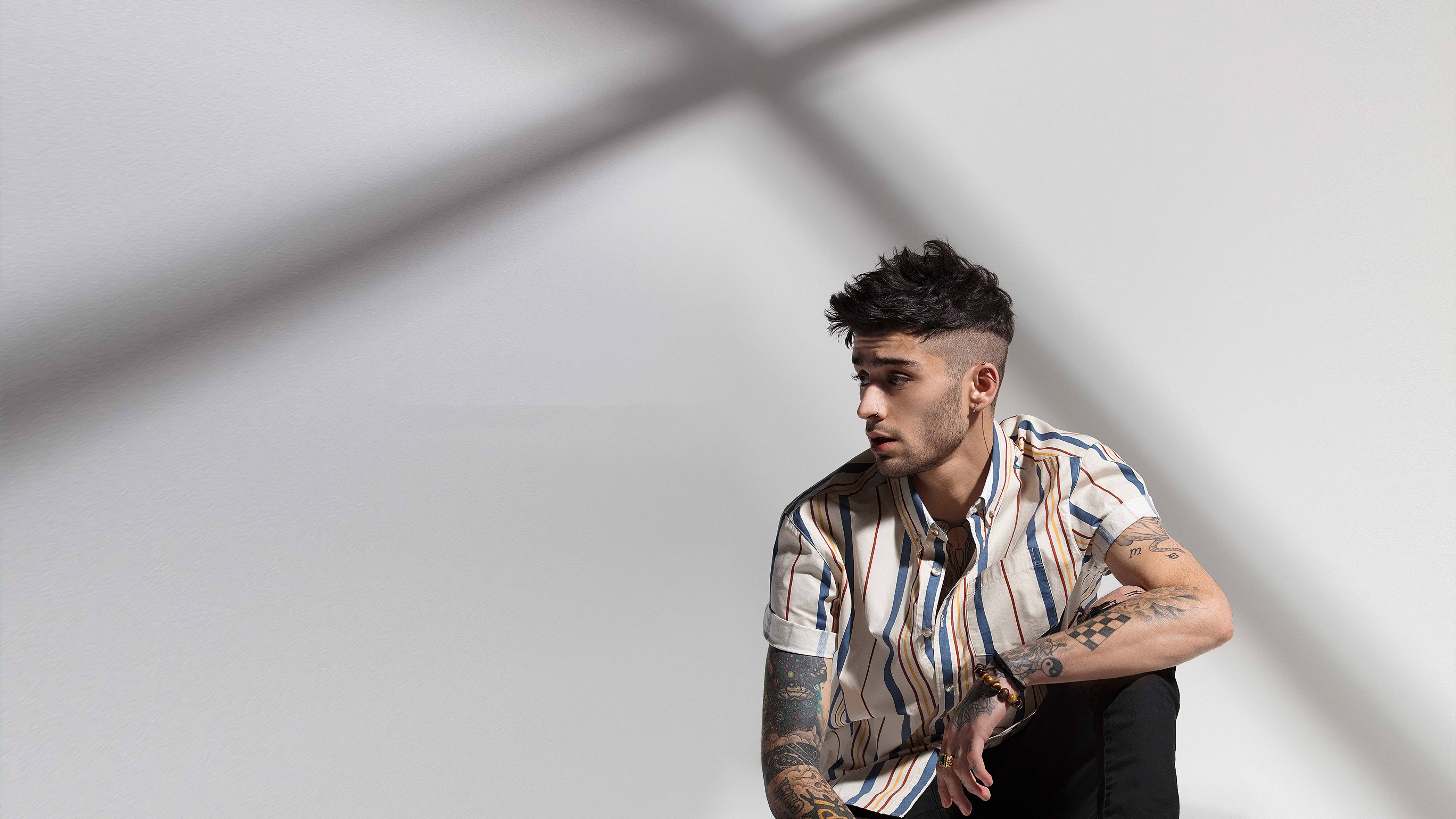 zayn 桌布 壁紙 — 一位有刺青和造型頭髮的年輕男子靠著白牆坐著，牆上有戲劇性的交叉陰影。
