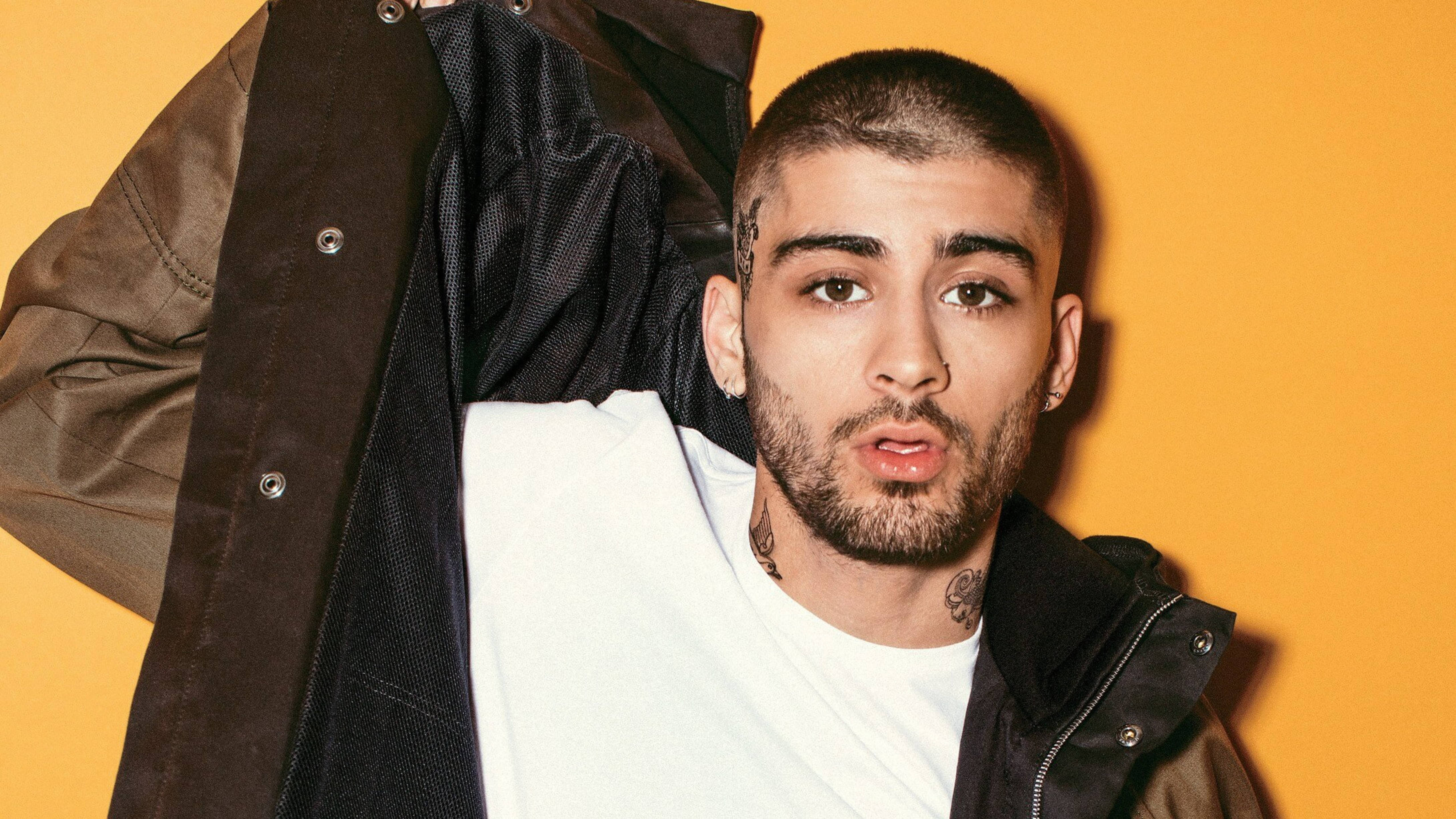 zayn 桌布 壁紙 — 一位有刺青和鼻環的男子，身穿棕色外套內搭白色T恤，在充滿活力的橙色背景前自信地擺姿勢。