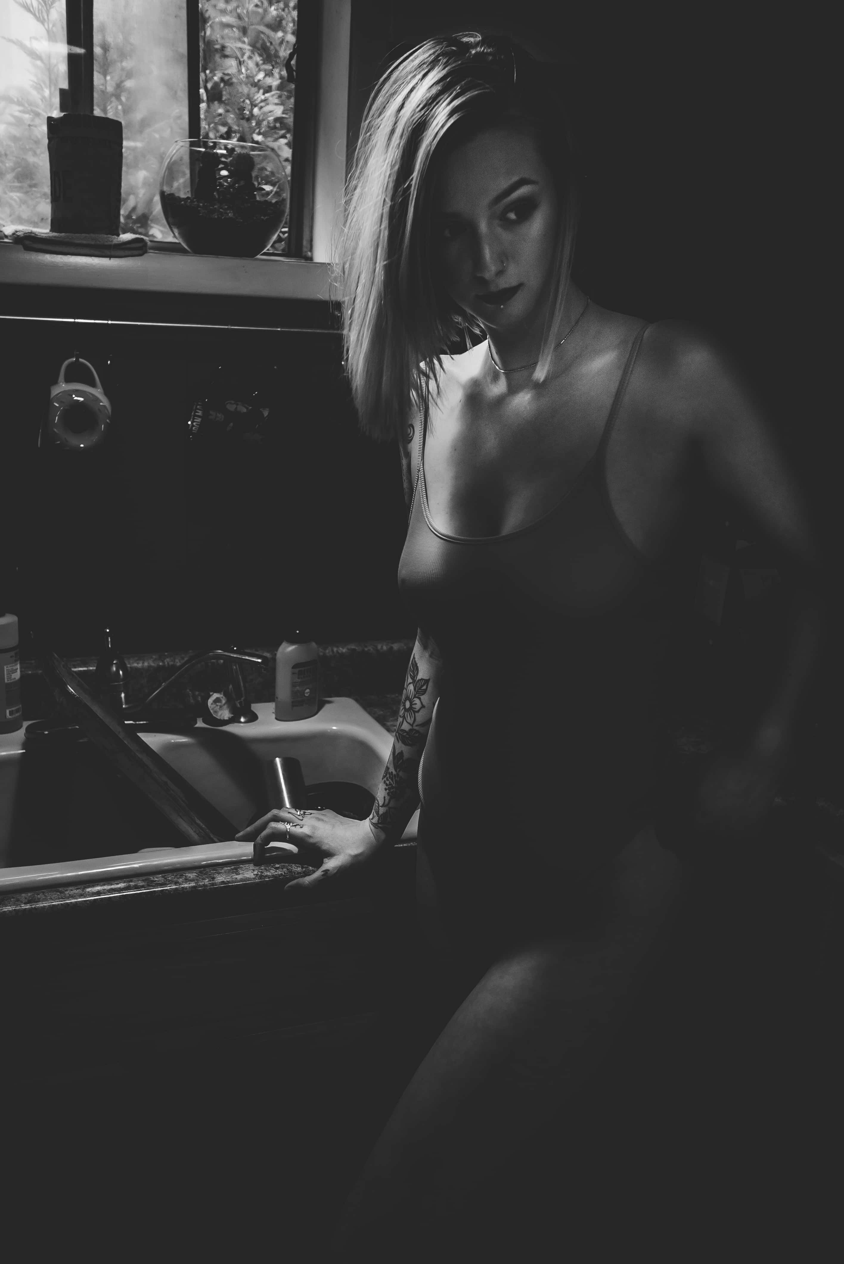 Sexy 桌布 壁紙 — 一位有刺青手臂的女人靠在昏暗廚房的洗手台旁，沐浴在柔和窗光中。