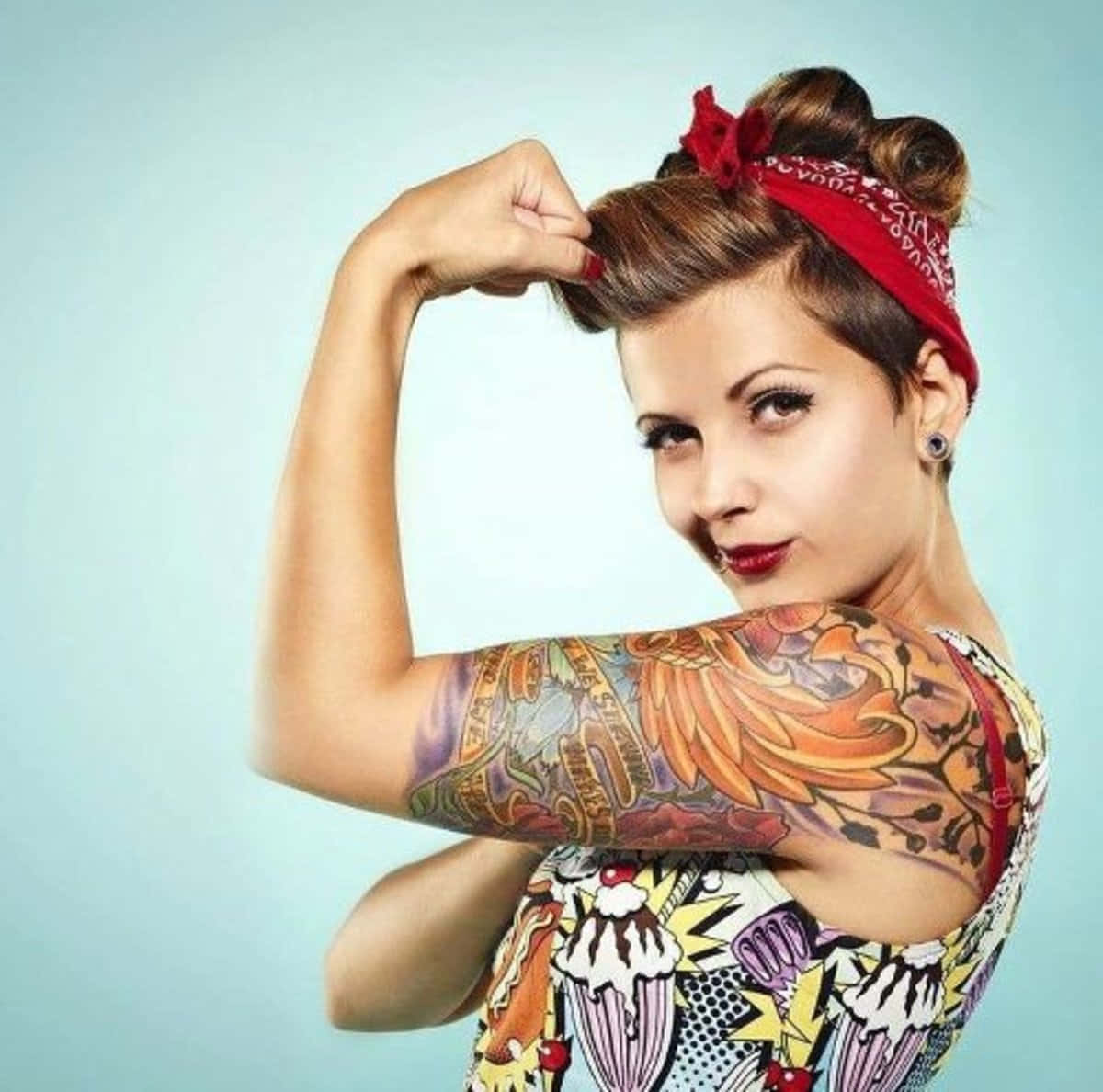 Tattoo Girl 桌布 壁紙 — 一位有刺青手臂的女性，戴著紅色頭巾，穿著普普藝術連衣裙，在淺藍色背景前彎曲二頭肌。