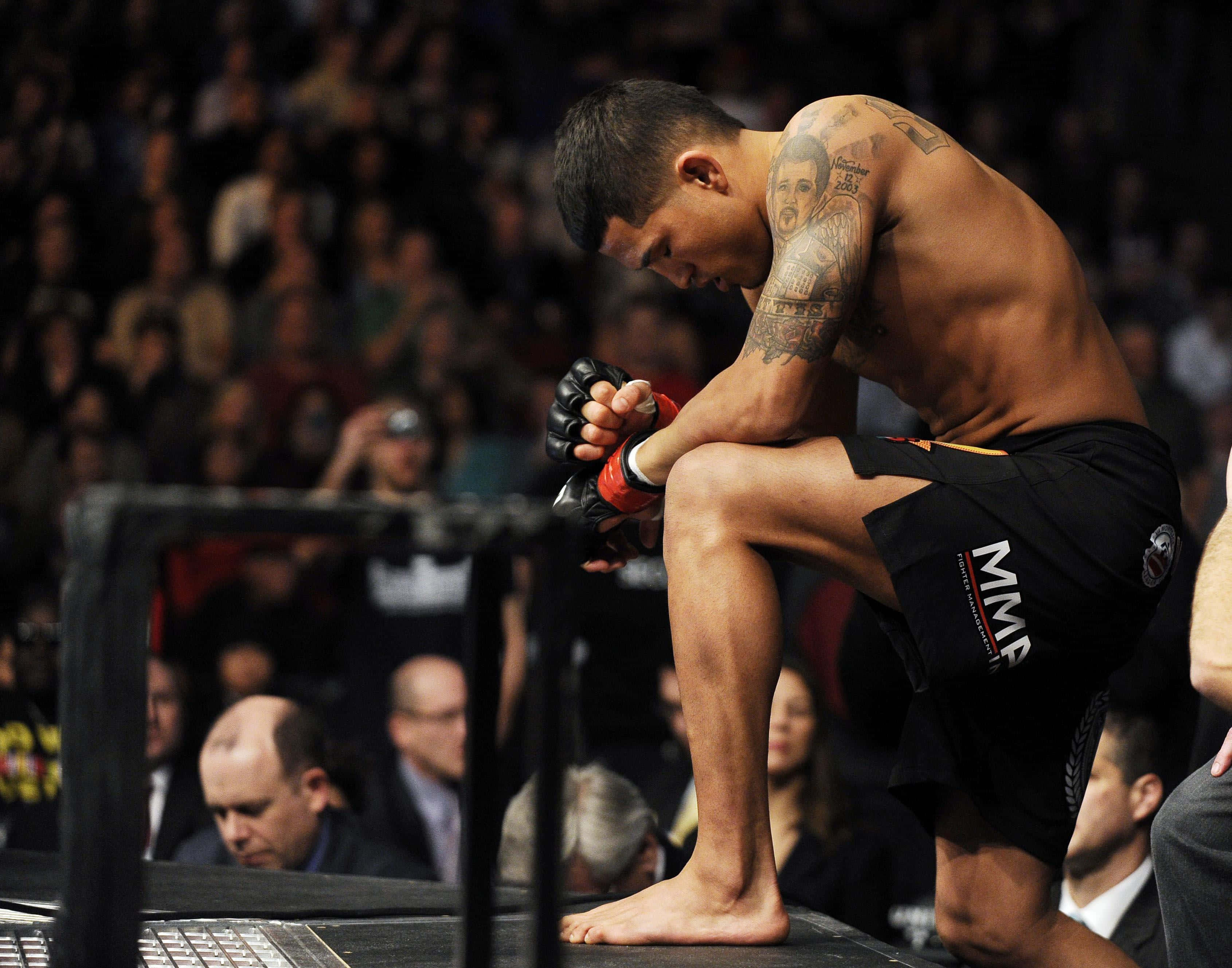UFC 桌布 壁紙 — 一位有刺青的MMA拳手坐在昏暗場地的籠邊，低頭合十，周圍是模糊的觀眾。