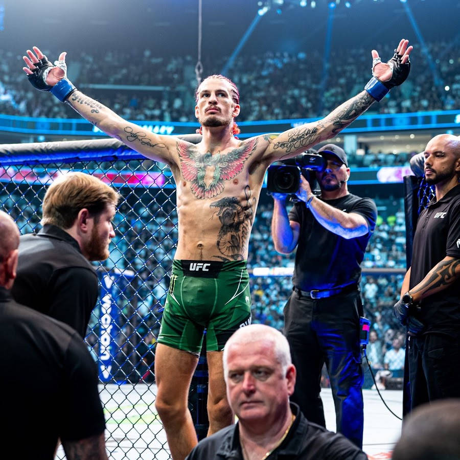 UFC 桌布 壁紙 — 一位有刺青的MMA拳擊手穿著綠色短褲，在燈光明亮的八角籠內舉起雙臂慶祝勝利，鐵絲網後方有歡呼的人群。
