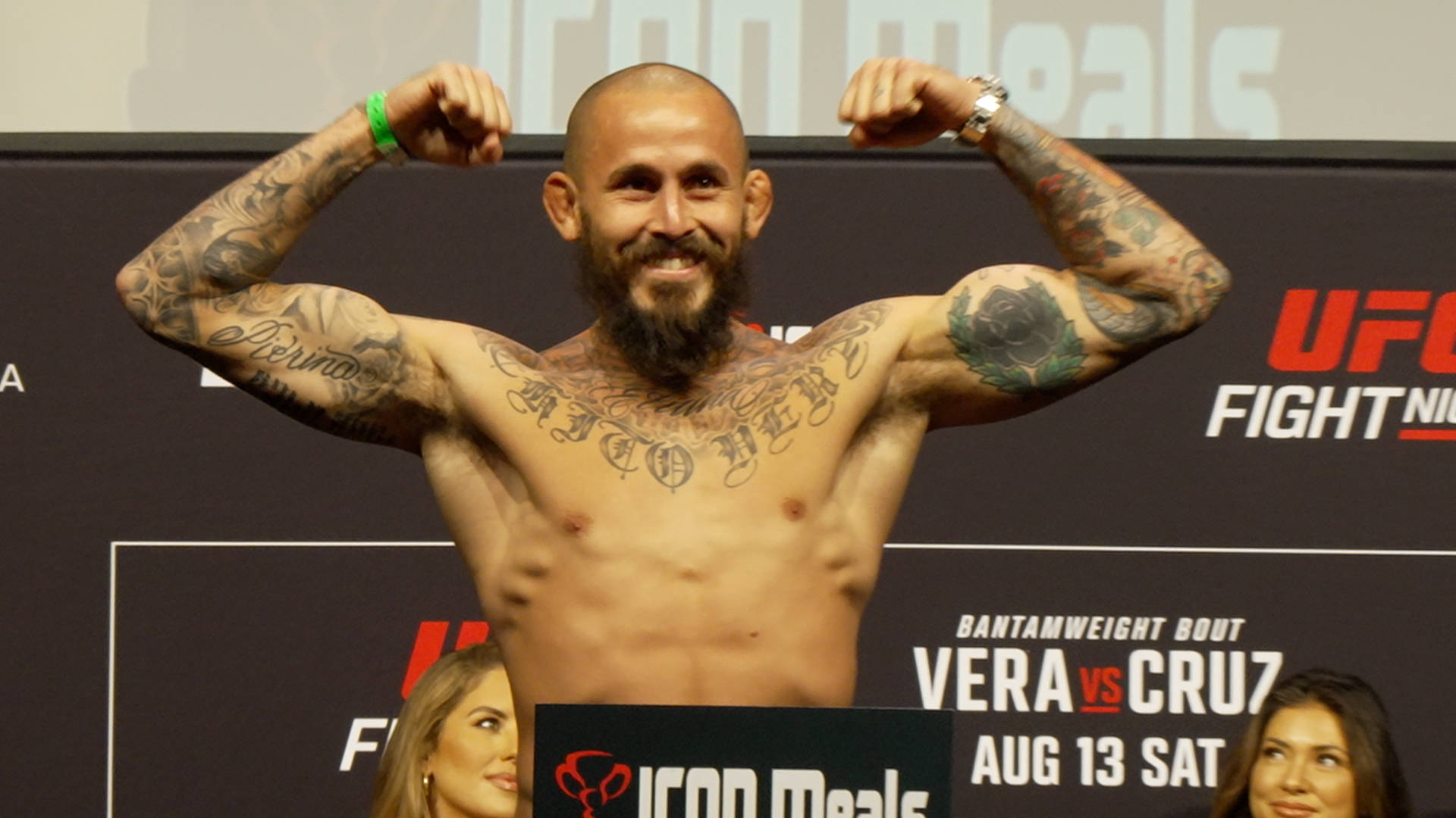 UFC 桌布 壁紙 — 一位有刺青的UFC拳手在明亮燈光下的稱重活動中咧嘴笑著展示肌肉。