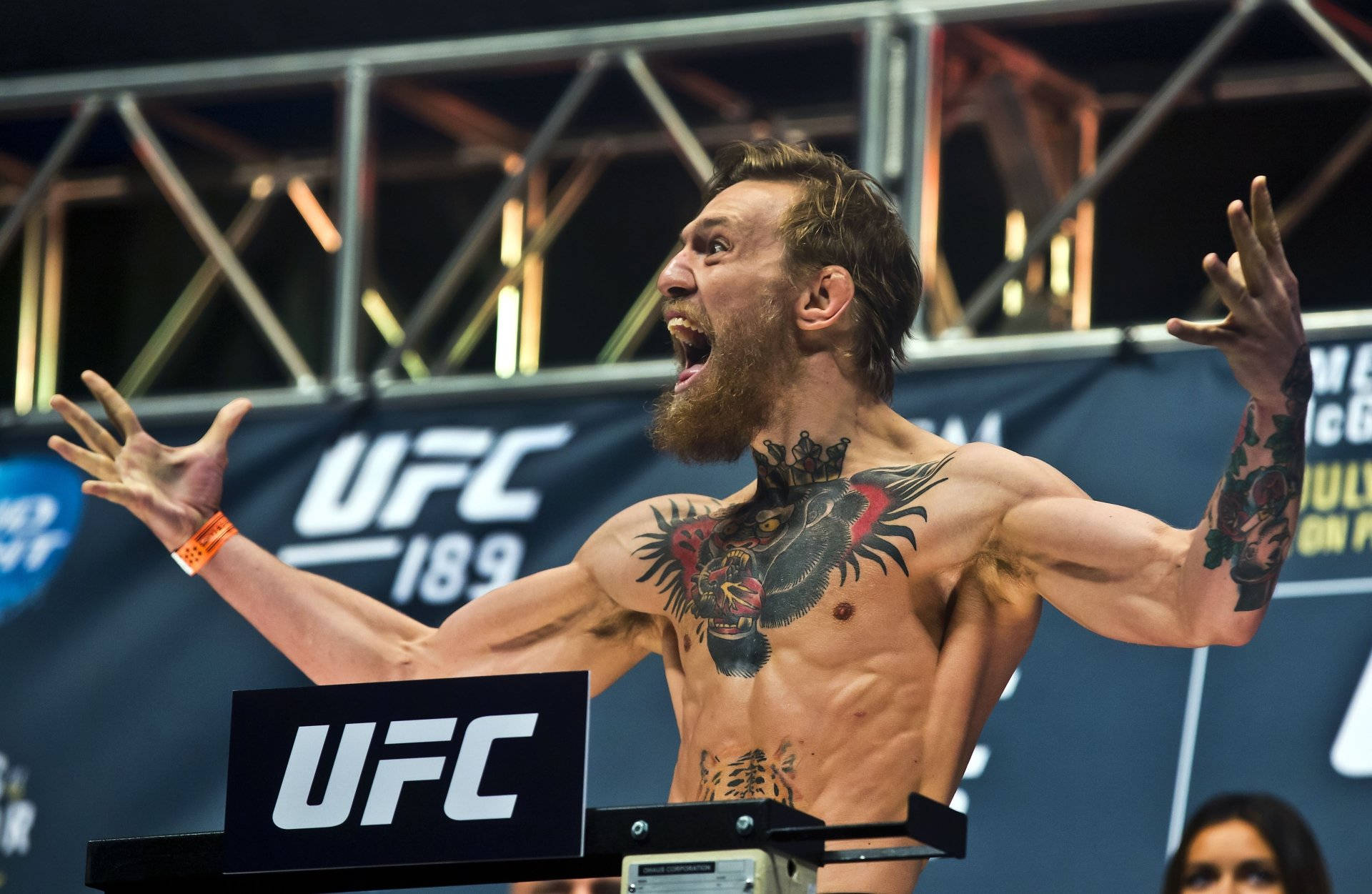 UFC 桌布 壁紙 — 一位有刺青的UFC拳手在明亮的場館燈光下，於過磅活動中張開雙臂，發出勝利的咆哮。