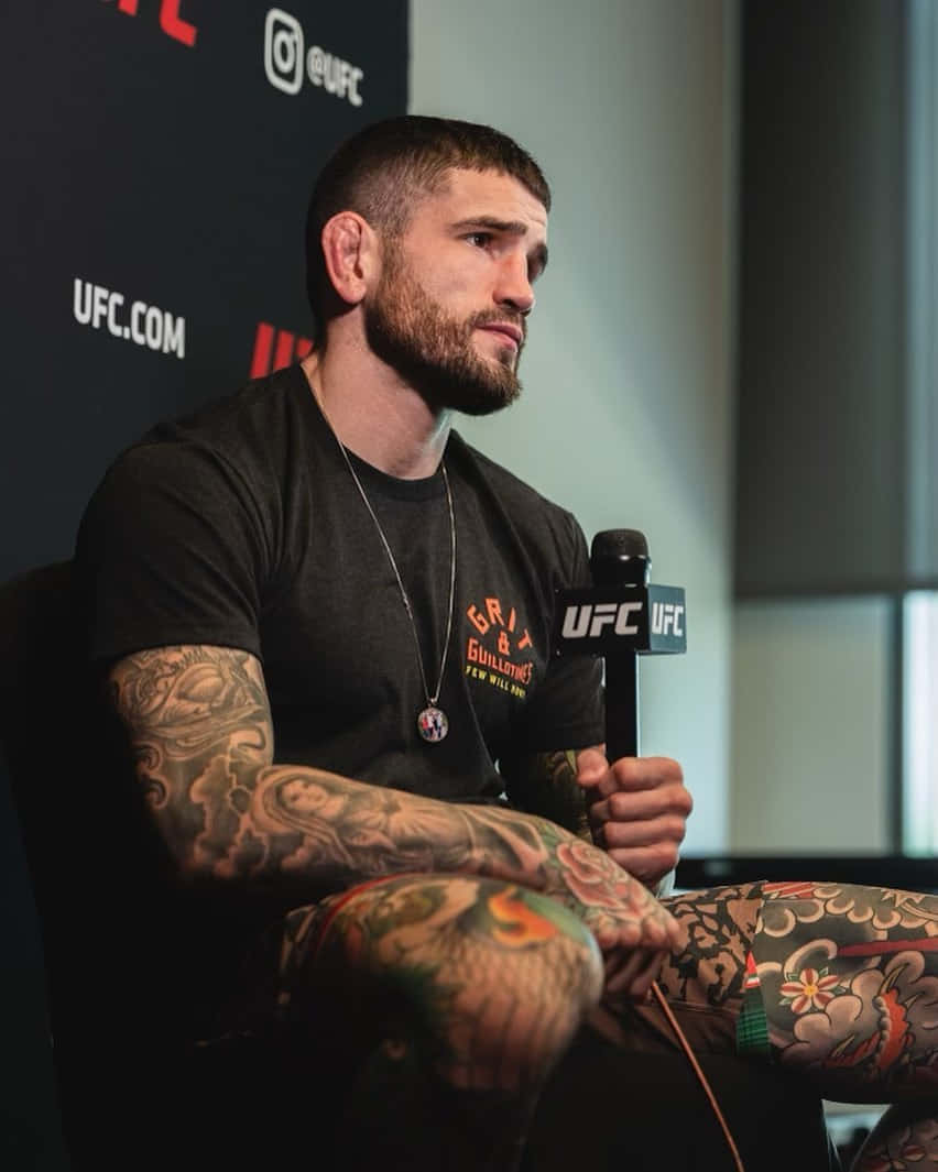 MMA 桌布 壁紙 — 一位有刺青的UFC拳手坐在記者會上，手持麥克風，表情嚴肅。