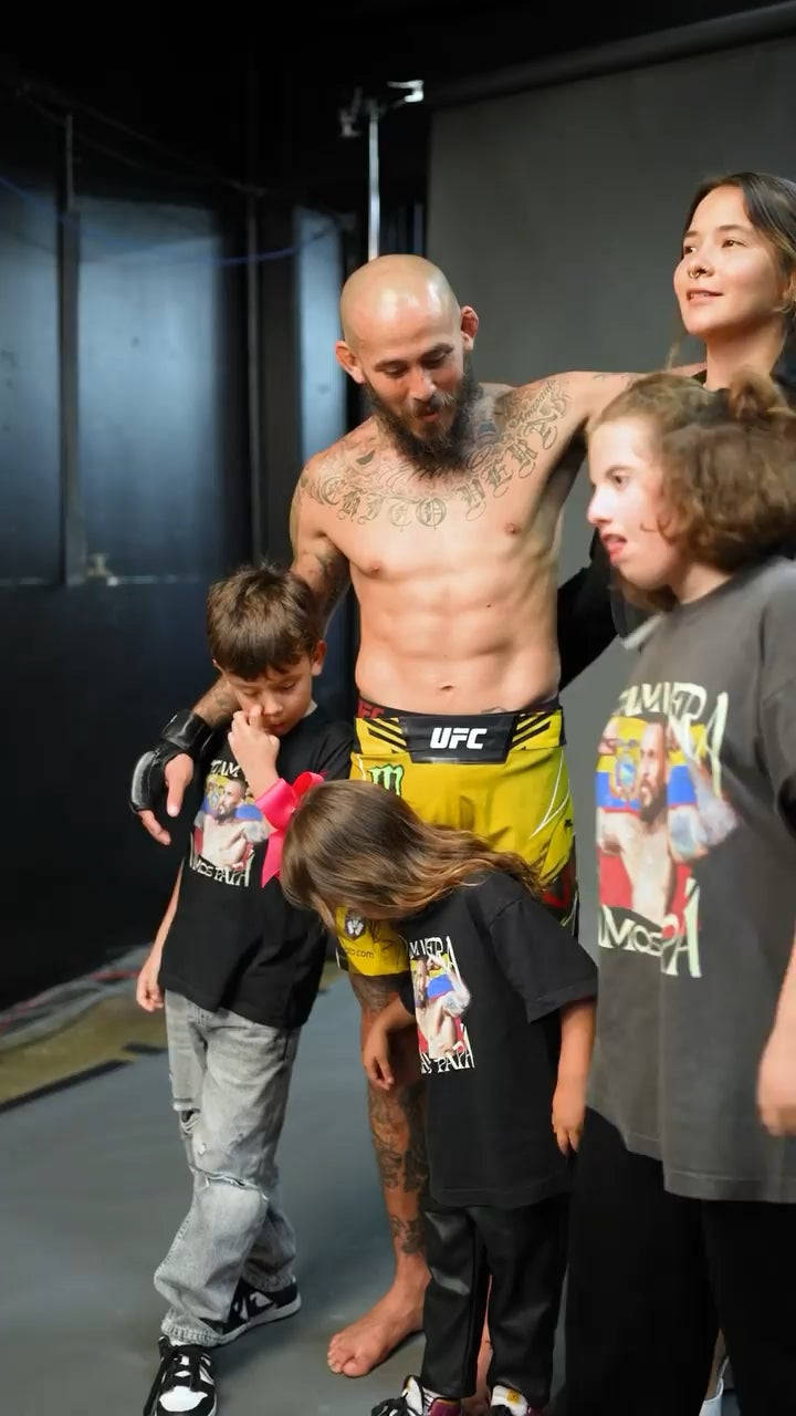 UFC 桌布 壁紙 — 一位有刺青的UFC拳手穿著黃色短褲，赤腳與四個孩子和一個女人站在昏暗的攝影棚中。
