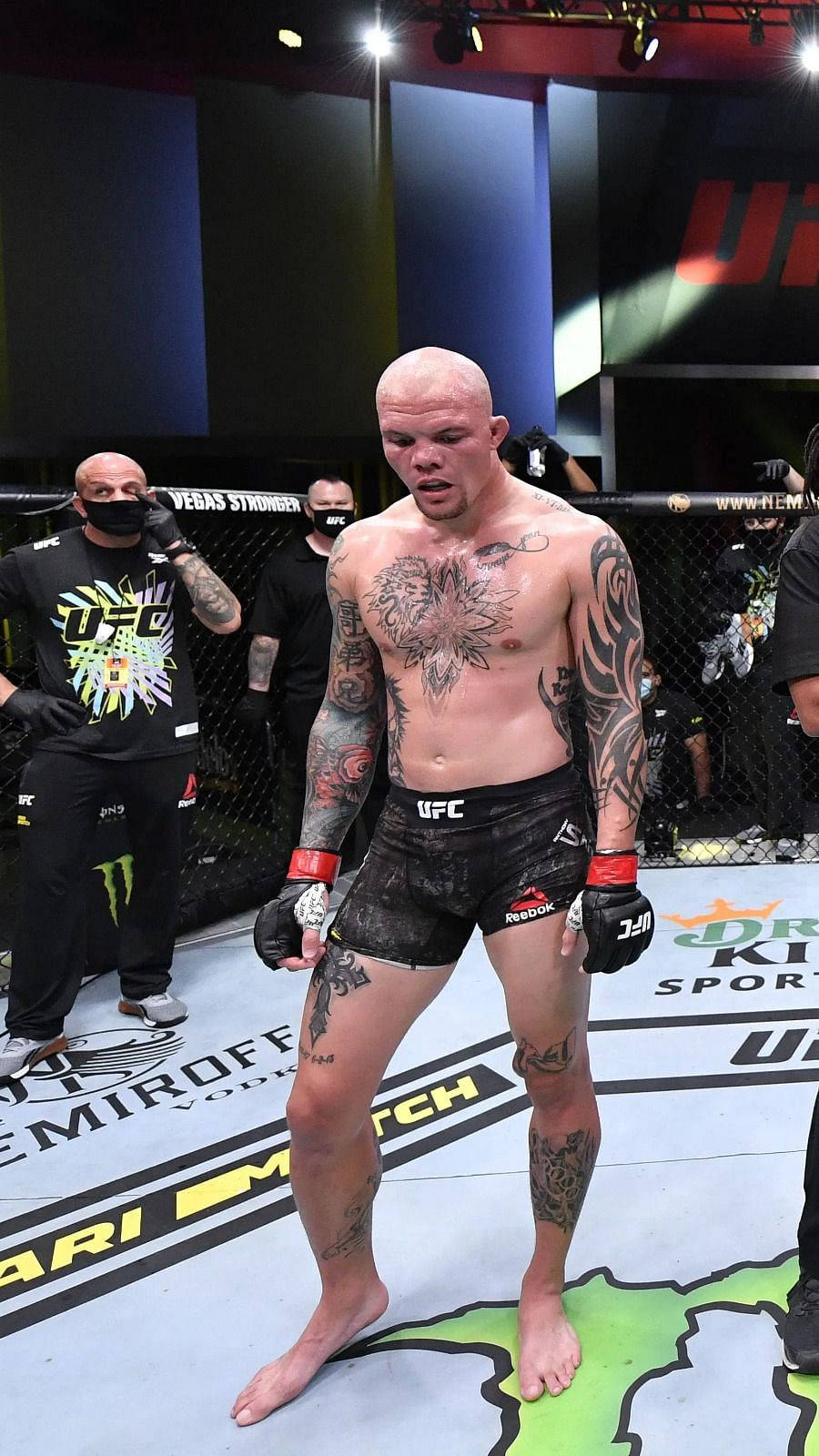 MMA 桌布 壁紙 — 一位有刺青的UFC拳手赤腳站在八角籠內，背景有工作人員，場館燈光明亮。