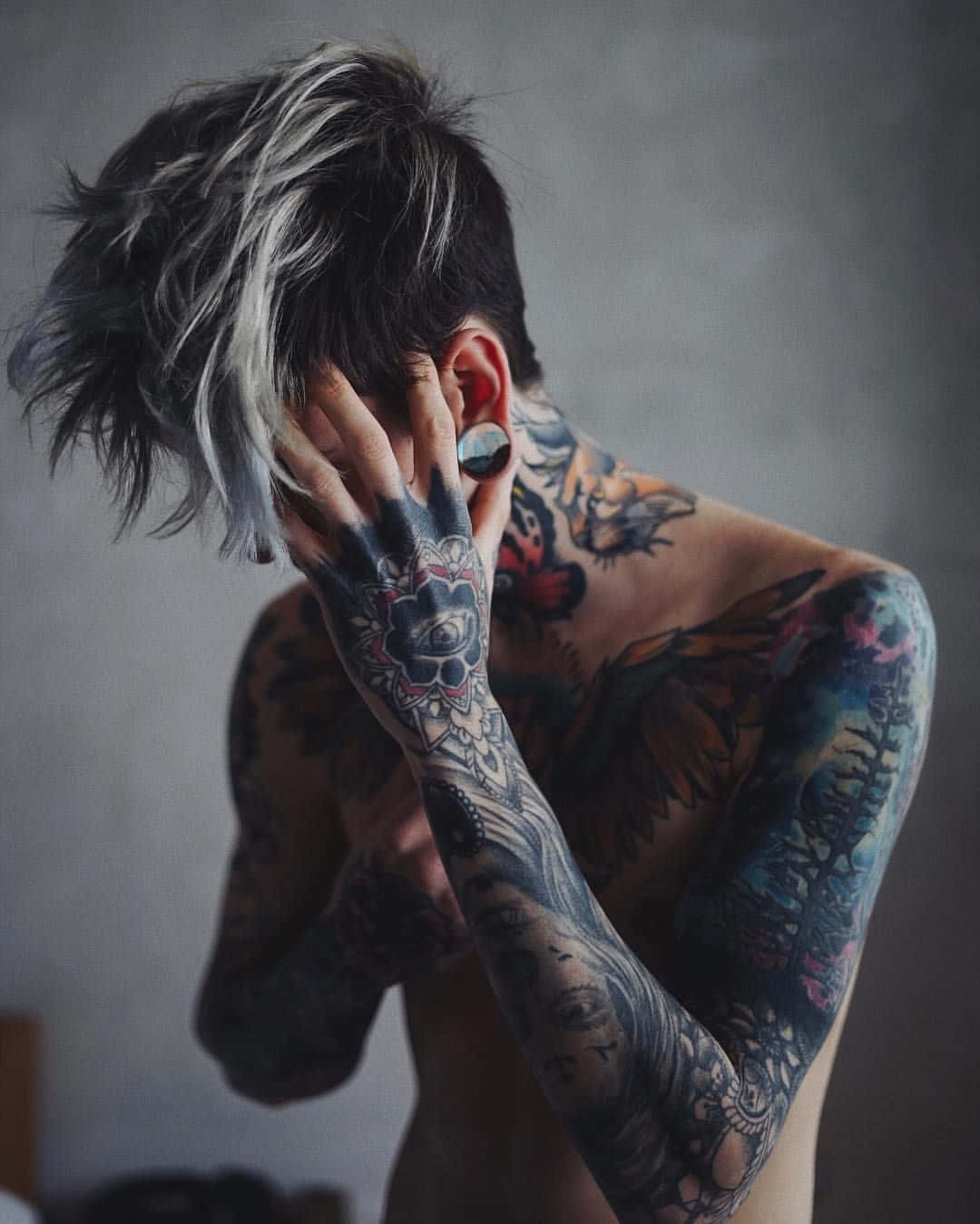 Tattoo Boy 桌布 壁紙 — 一位有刺青的人，銀色和黑色頭髮，用有刺青的手遮住臉，背景是柔和的灰色。