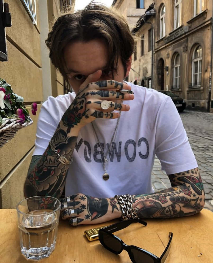 Tattoo Boy 桌布 壁紙 — 一位有刺青的人坐在狹窄歐洲街道的戶外咖啡桌旁，用手遮住半邊臉。