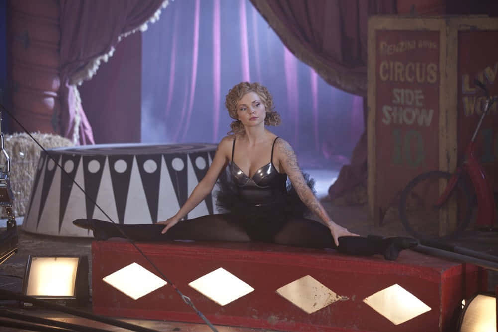 Izabella Miko 桌布 壁紙 — 一位有刺青的女士優雅地坐在紅色馬戲團平台上，紫色窗簾下，身後有「Circus Side Show」標誌。