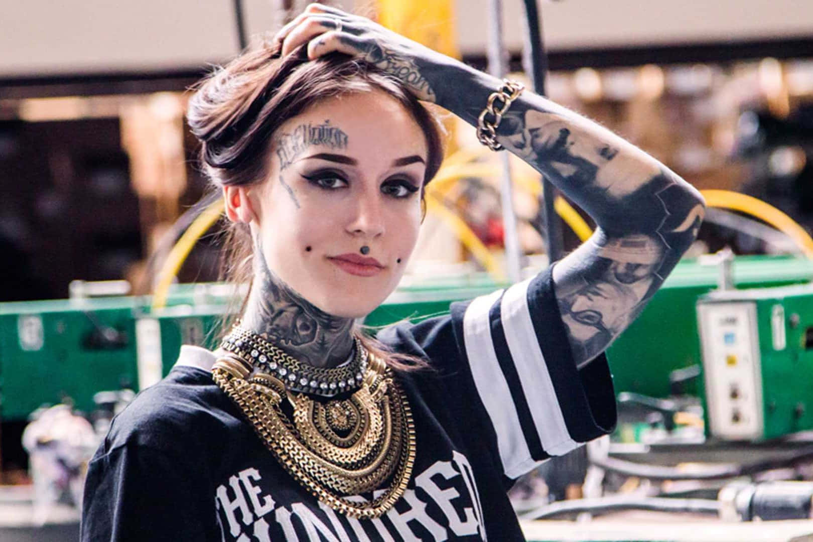 Tattoo Girl 桌布 壁紙 — 一位有刺青的女性，佩戴金飾和黑色球衣，在工作室環境中自信地擺姿勢。