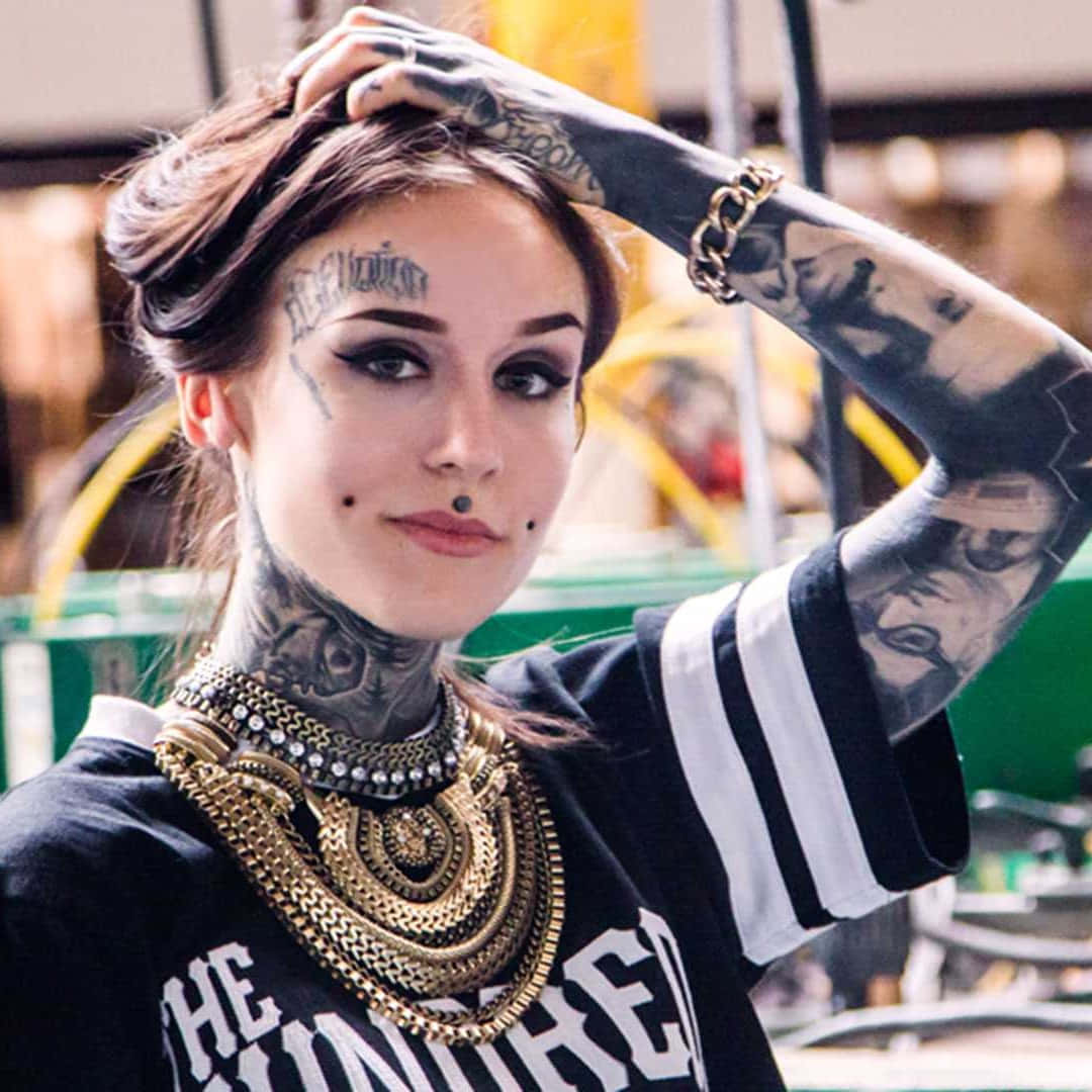 Tattoo Girl 桌布 壁紙 — 一位有刺青的女性，化著大膽的妝容，戴著多層金鍊，自信地將一隻手放在頭髮中擺姿勢。