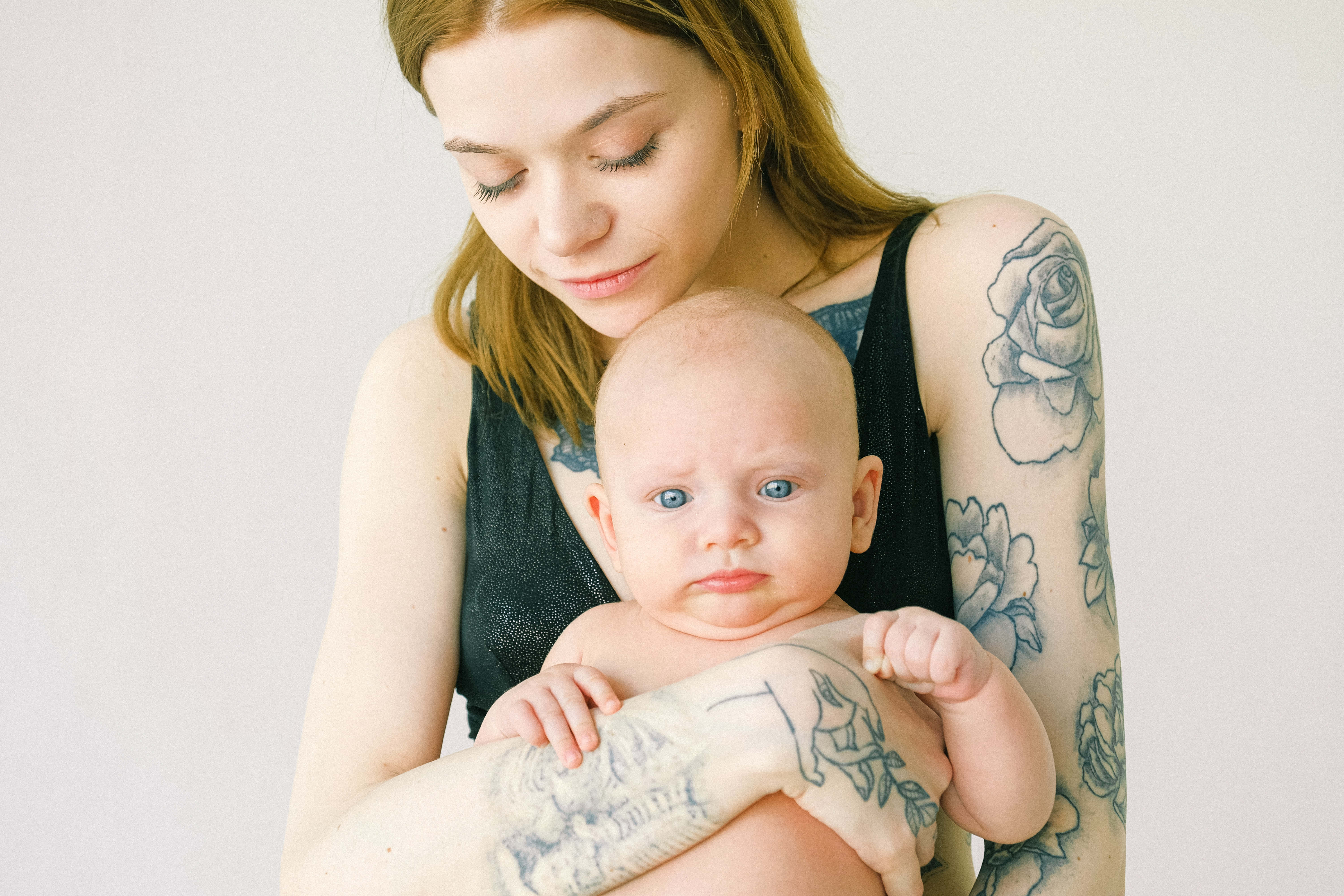 Mom 桌布 壁紙 — 一位有刺青的女性在白色背景前輕輕抱著一個擁有引人注目的藍色眼睛的嬰兒。