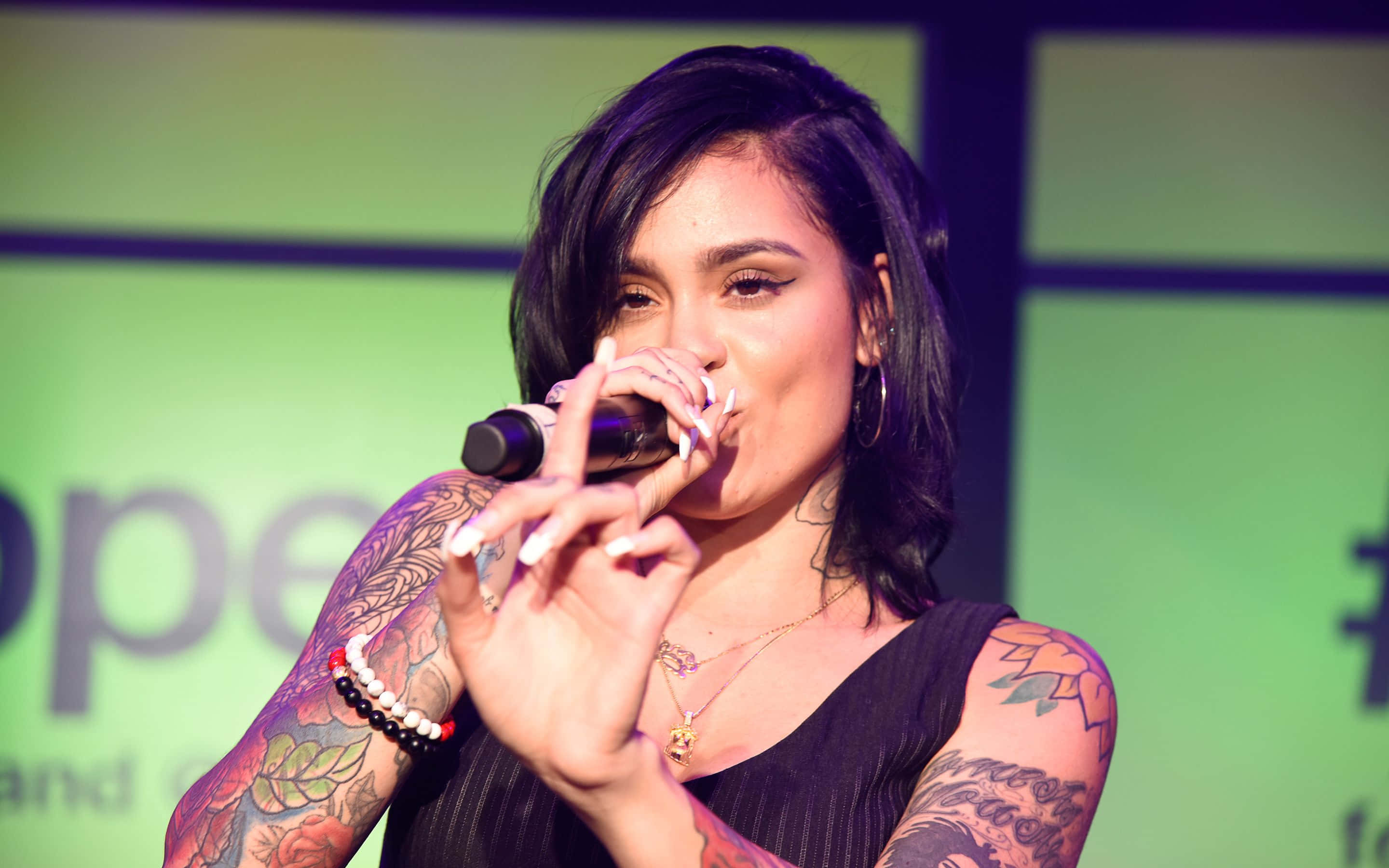 kehlani 桌布 壁紙 — 一位有刺青的女性在綠色背景的舞台燈光下熱情地對著麥克風唱歌。