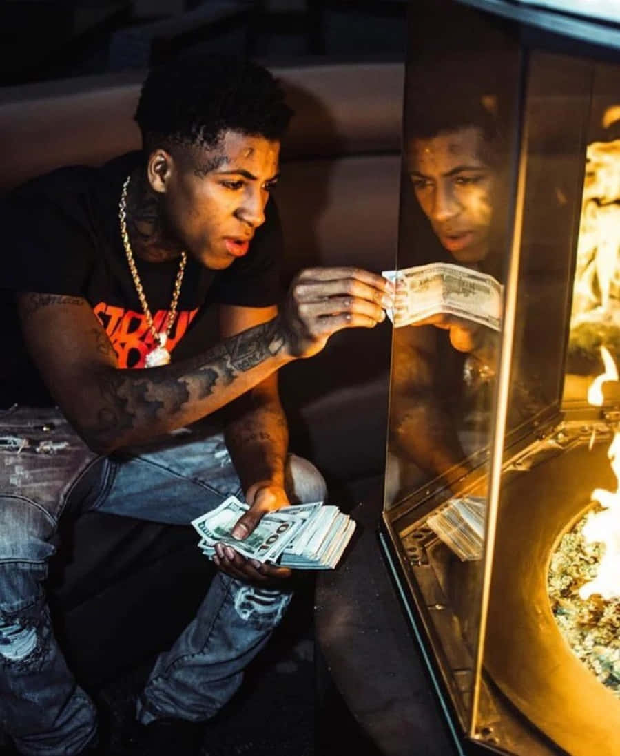 NBA YoungBoy 桌布 壁紙 — 一位有刺青的年輕男子在壁爐旁拿著現金，而他的倒影在玻璃中回望。