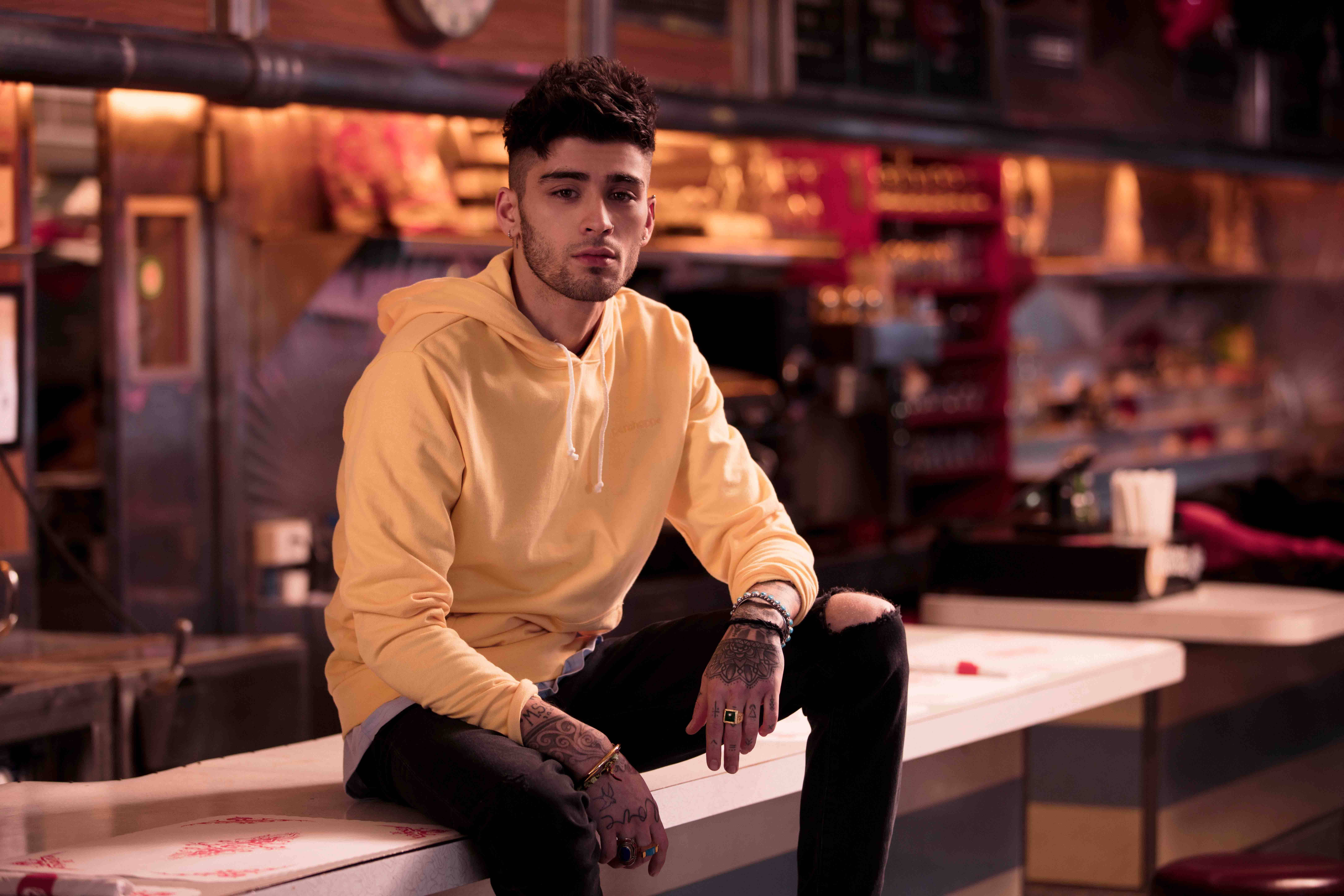 zayn 桌布 壁紙 — 一位有刺青的年輕男子在溫暖的環境燈光下，穿著黃色連帽衫，隨意地坐在餐廳櫃檯上。